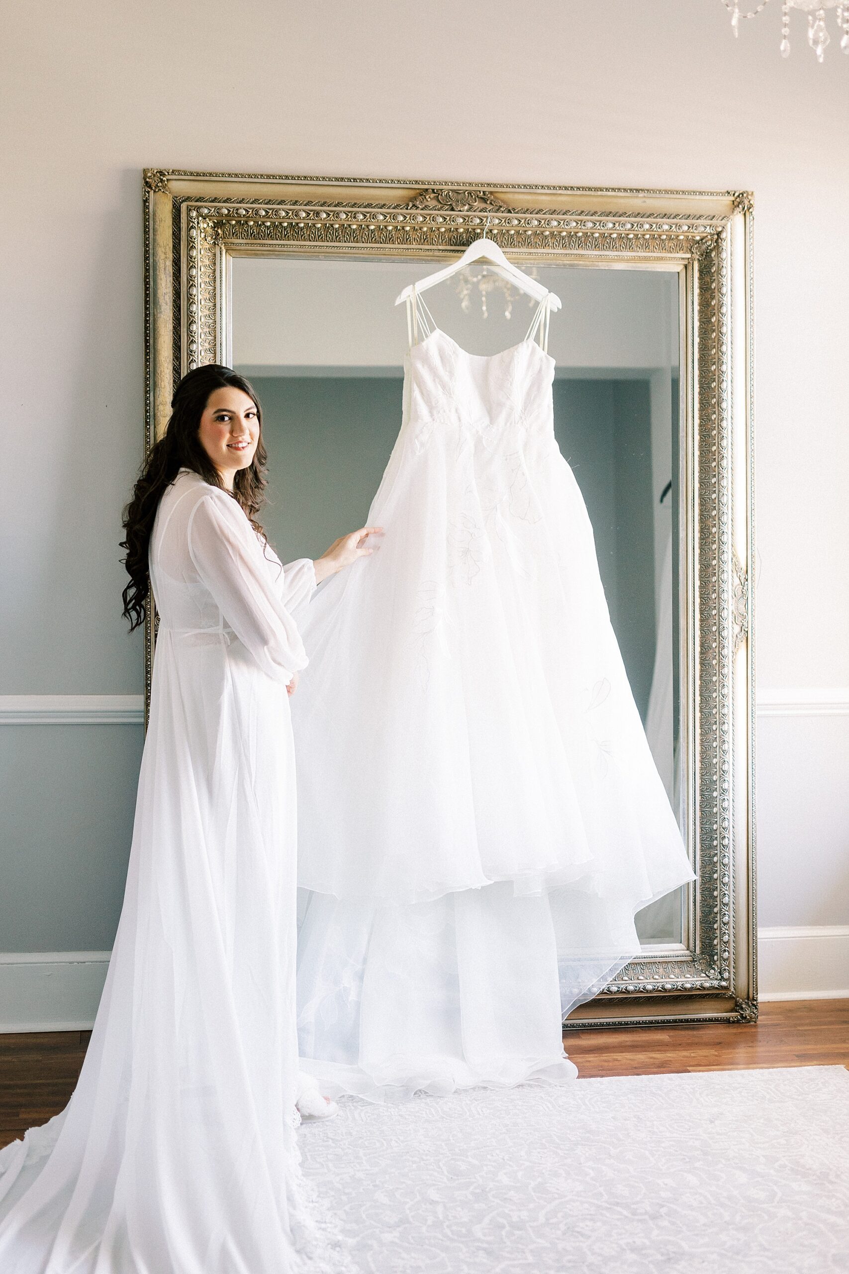 bride stands by mirror with wedding dress hanging on frame 
