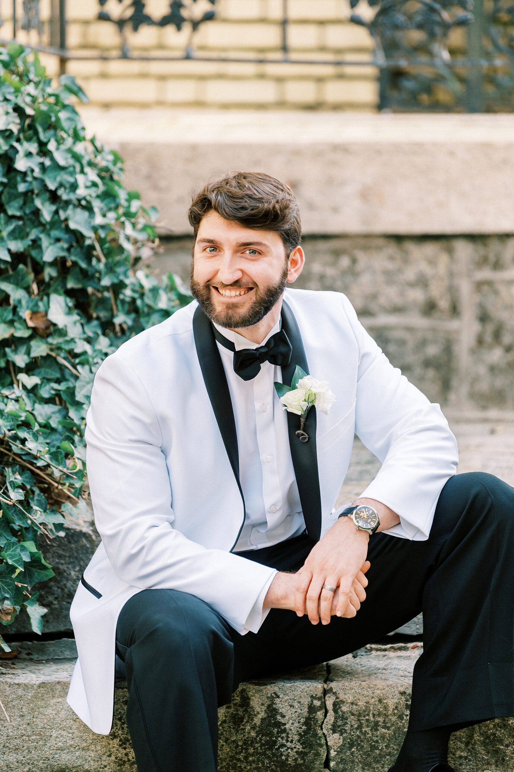 groom in white suit jacket sits on stone steps leaning into leg 
