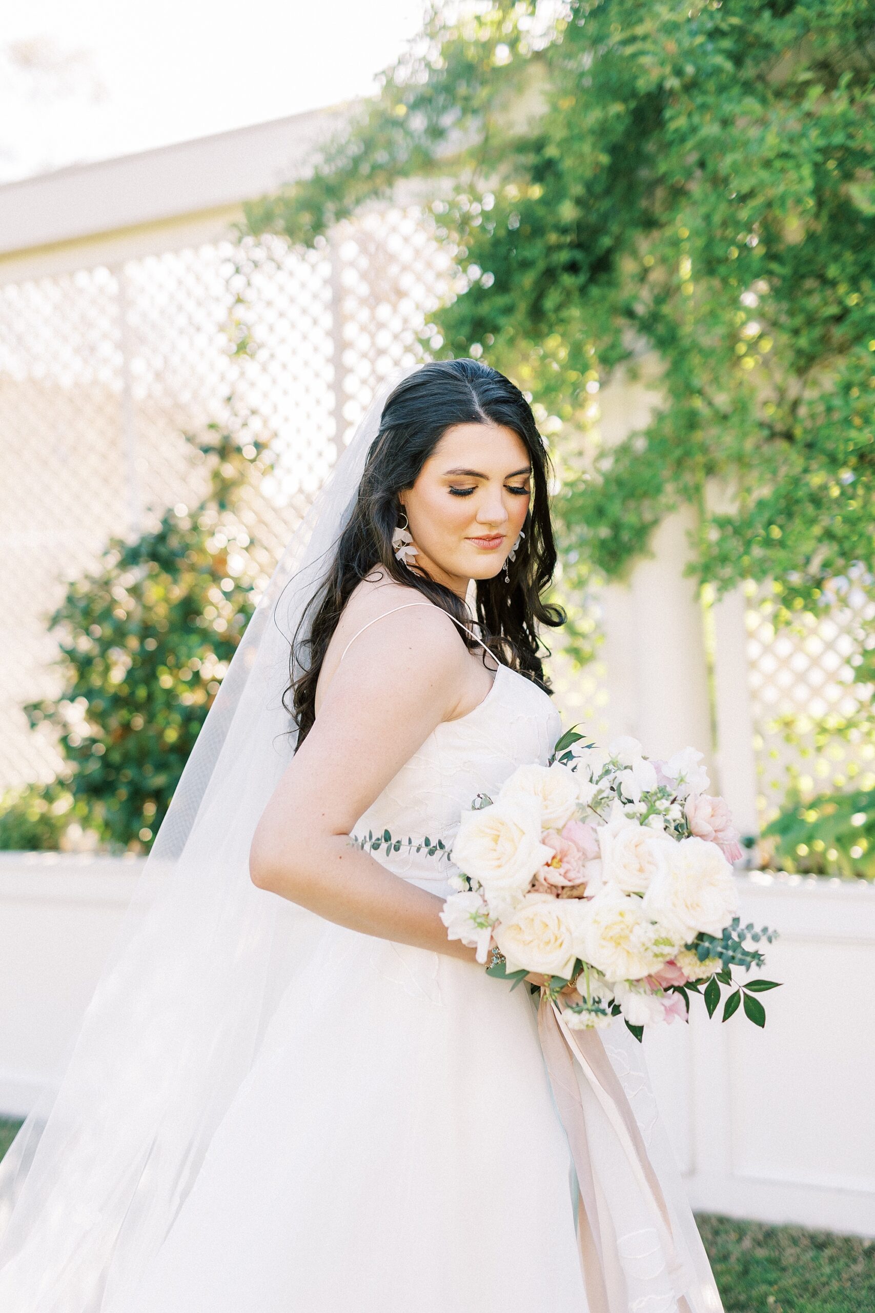 bride looks down at bouquet of pink and ivory flowers with veil behind her 
