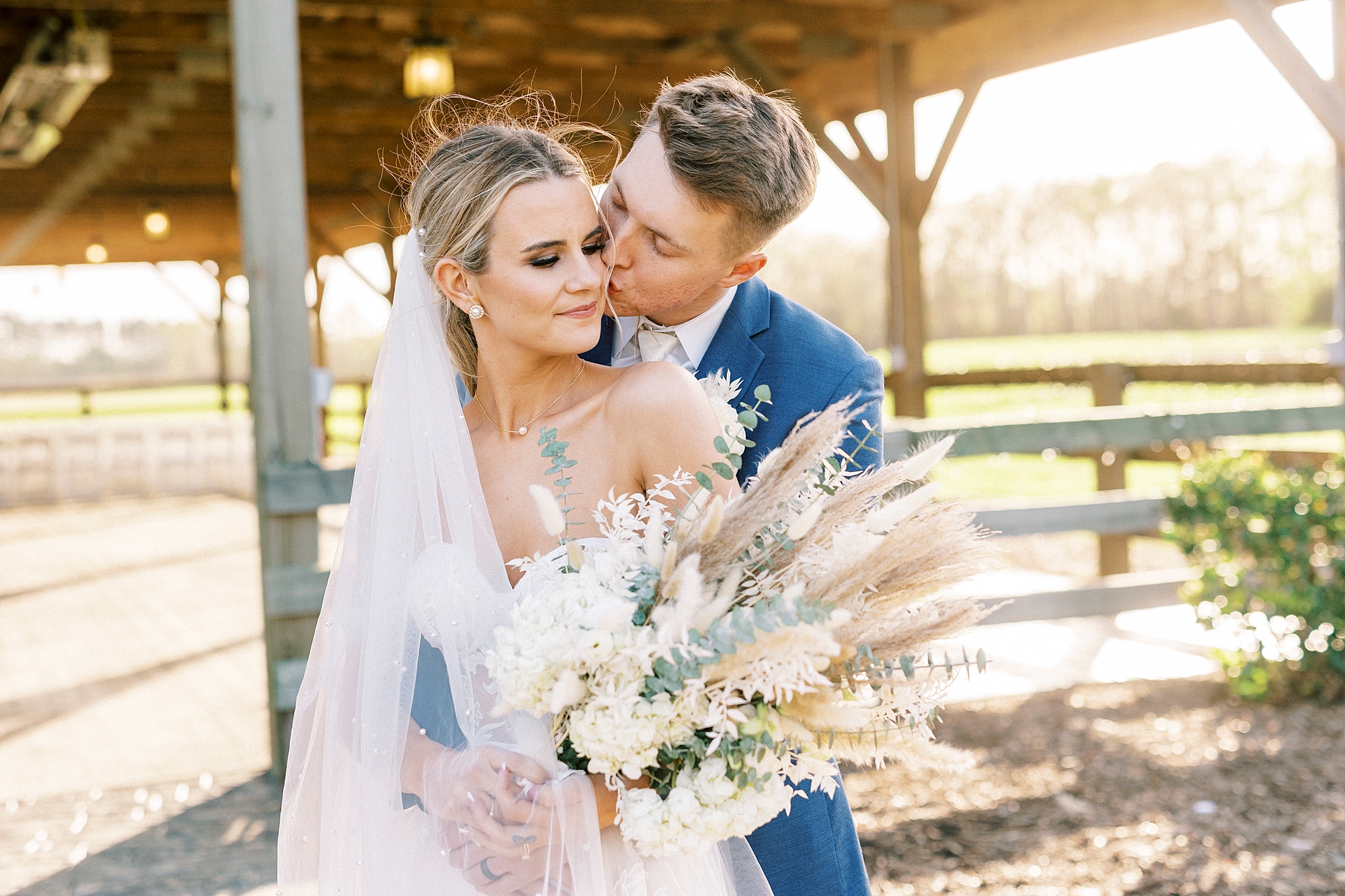 groom hugs bride from behind kissing her cheek with white flowers and Rhodes grass 