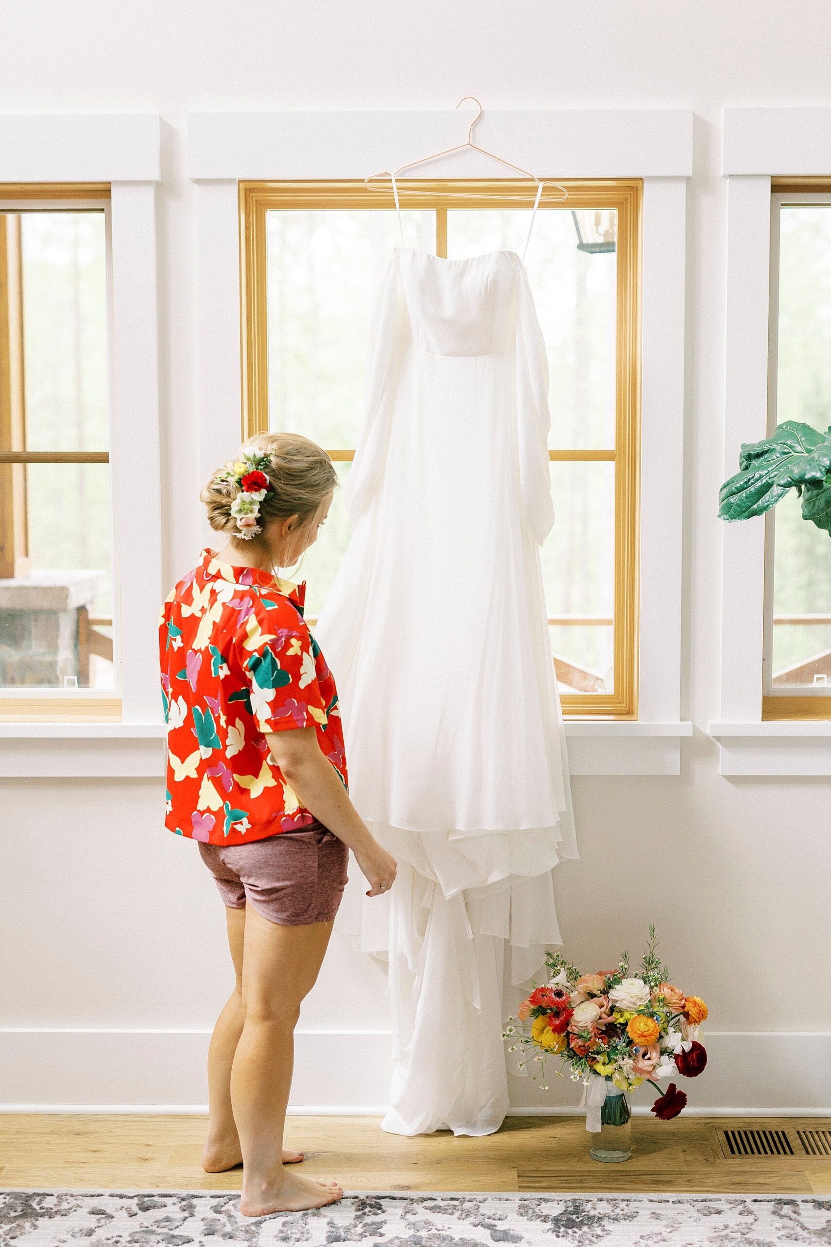 bride in red flower pajamas looks at wedding dress hanging in window 