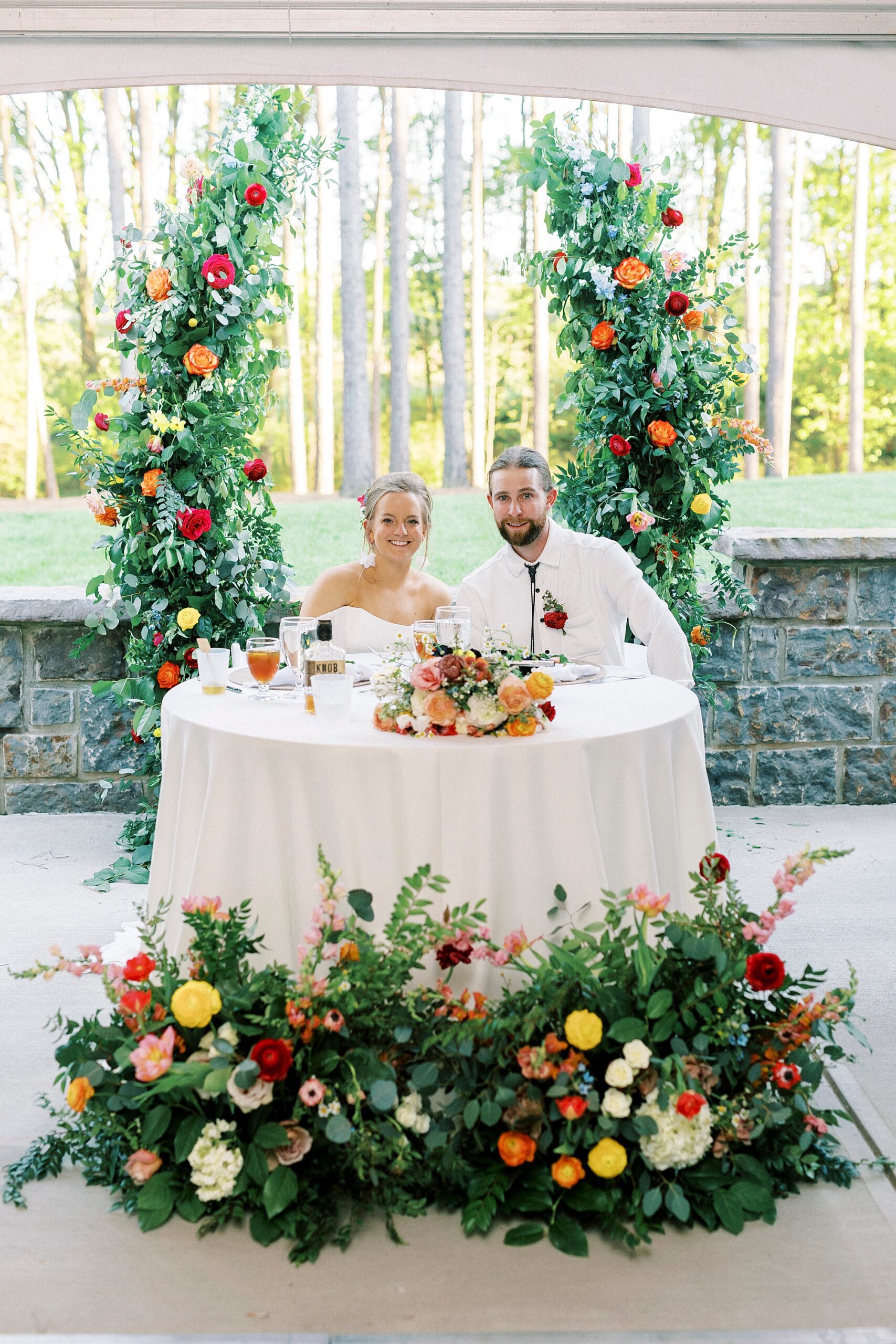 newlyweds sit at sweetheart table between three installations of red, pink, and yellow flowers 