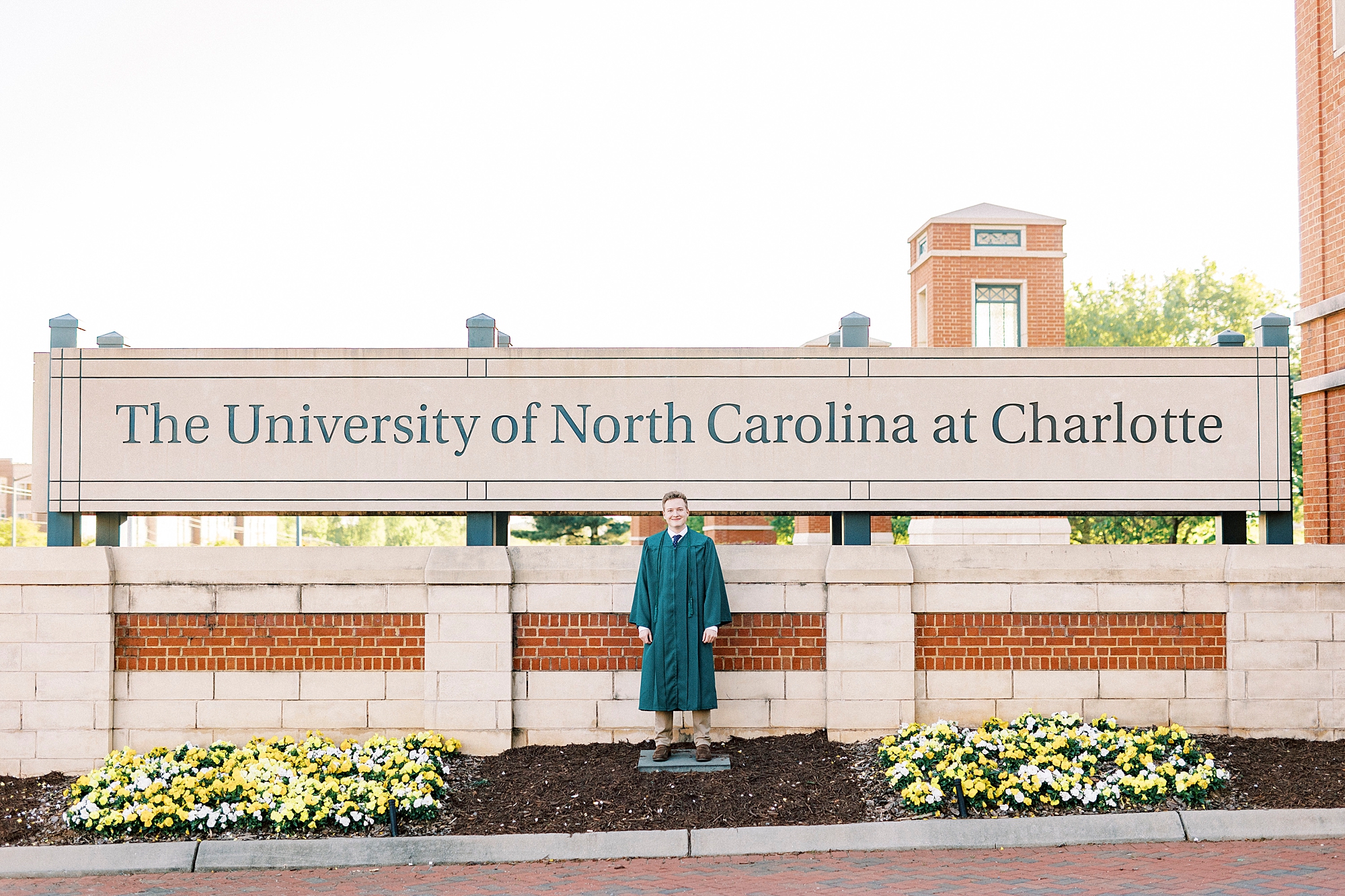 senior in green cap and gown poses by University of North Carolina at Charlotte sign