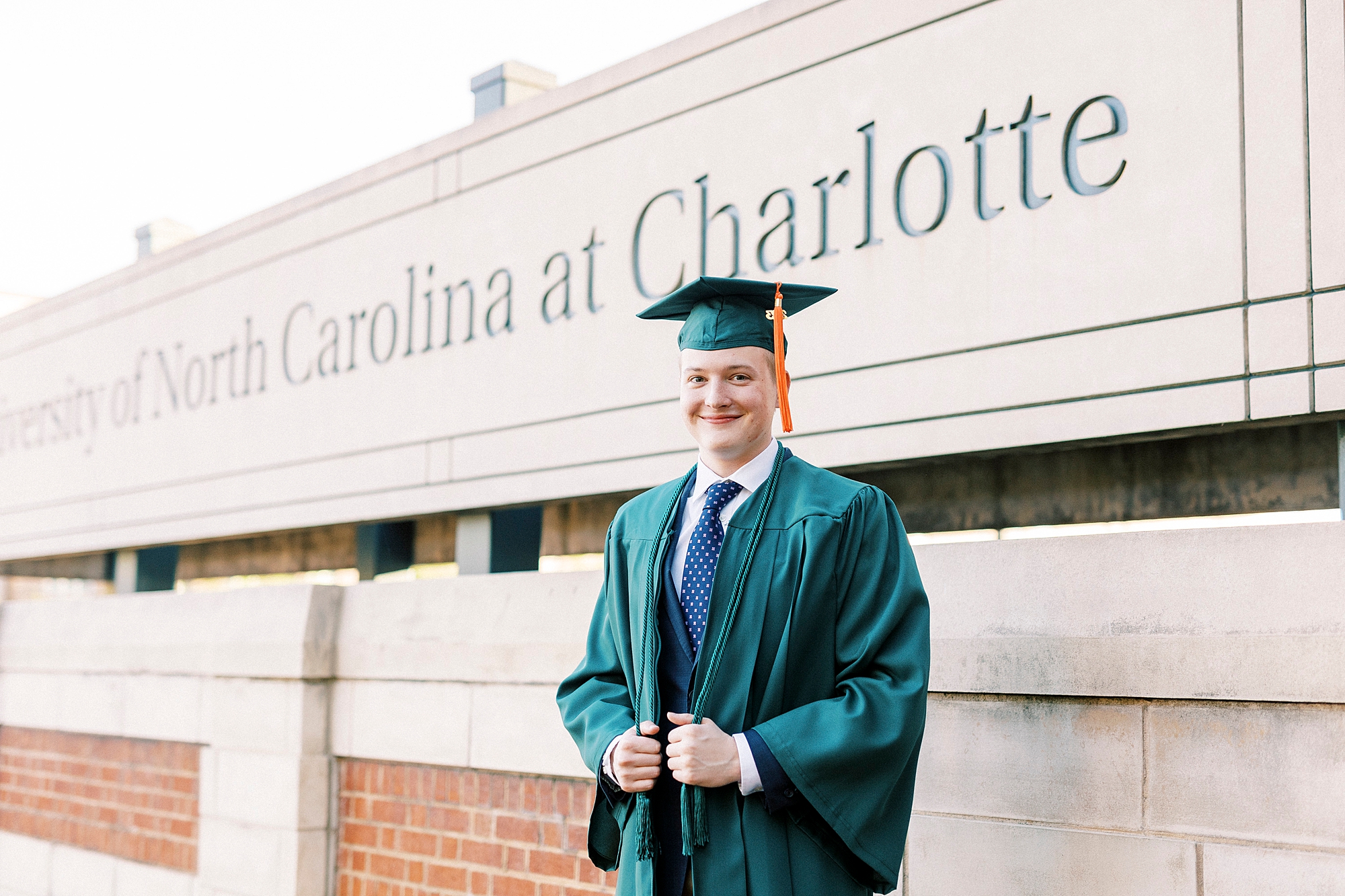 graduating senior in green cap and gown poses near University of North Carolina at Charlotte sign