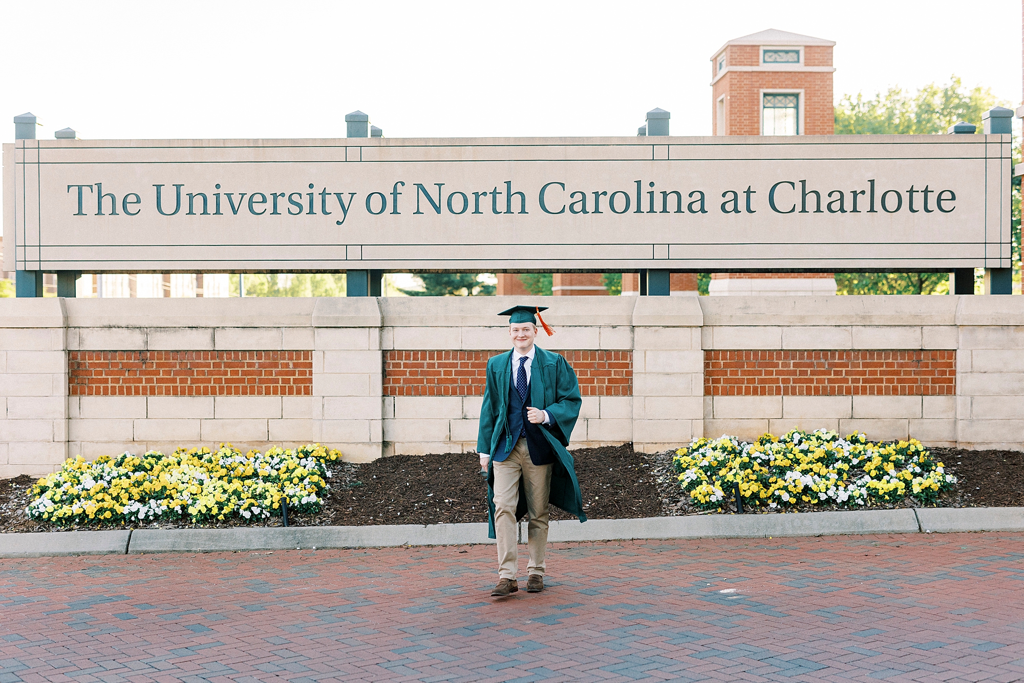 senior in green cap and gown walks in front of University of North Carolina at Charlotte sign