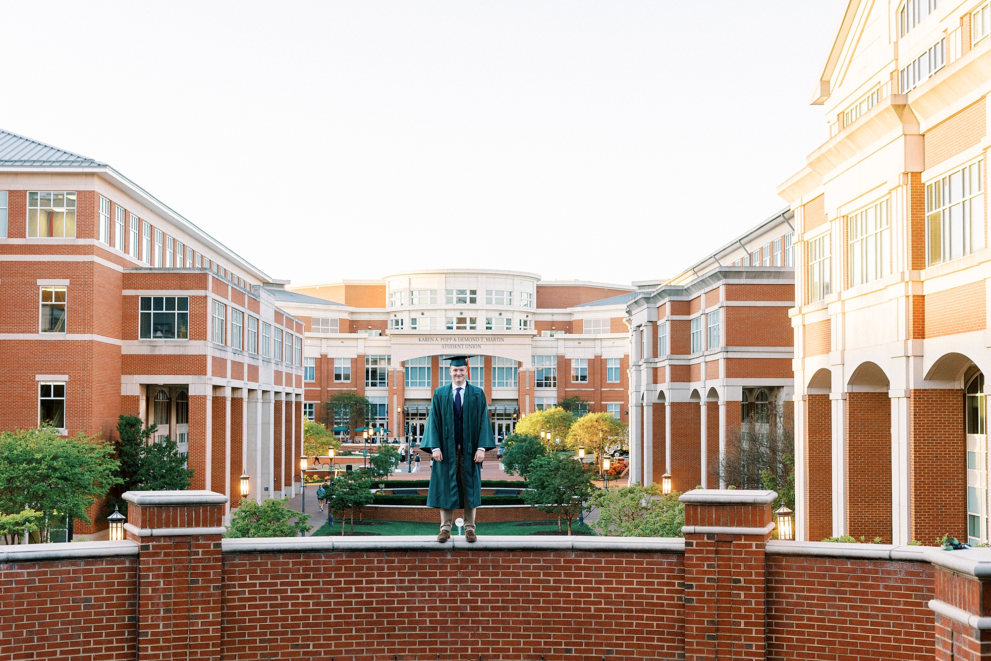 graduating senior in green cap and gown stands on brick wall during UNC Charlotte graduation portraits 