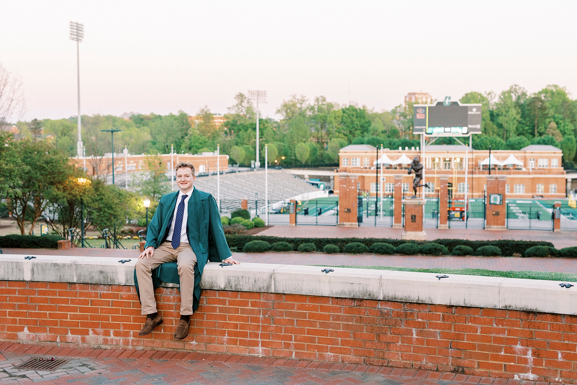 graduating senior in green gown sits on brick wall looking over UNC Charlotte football field