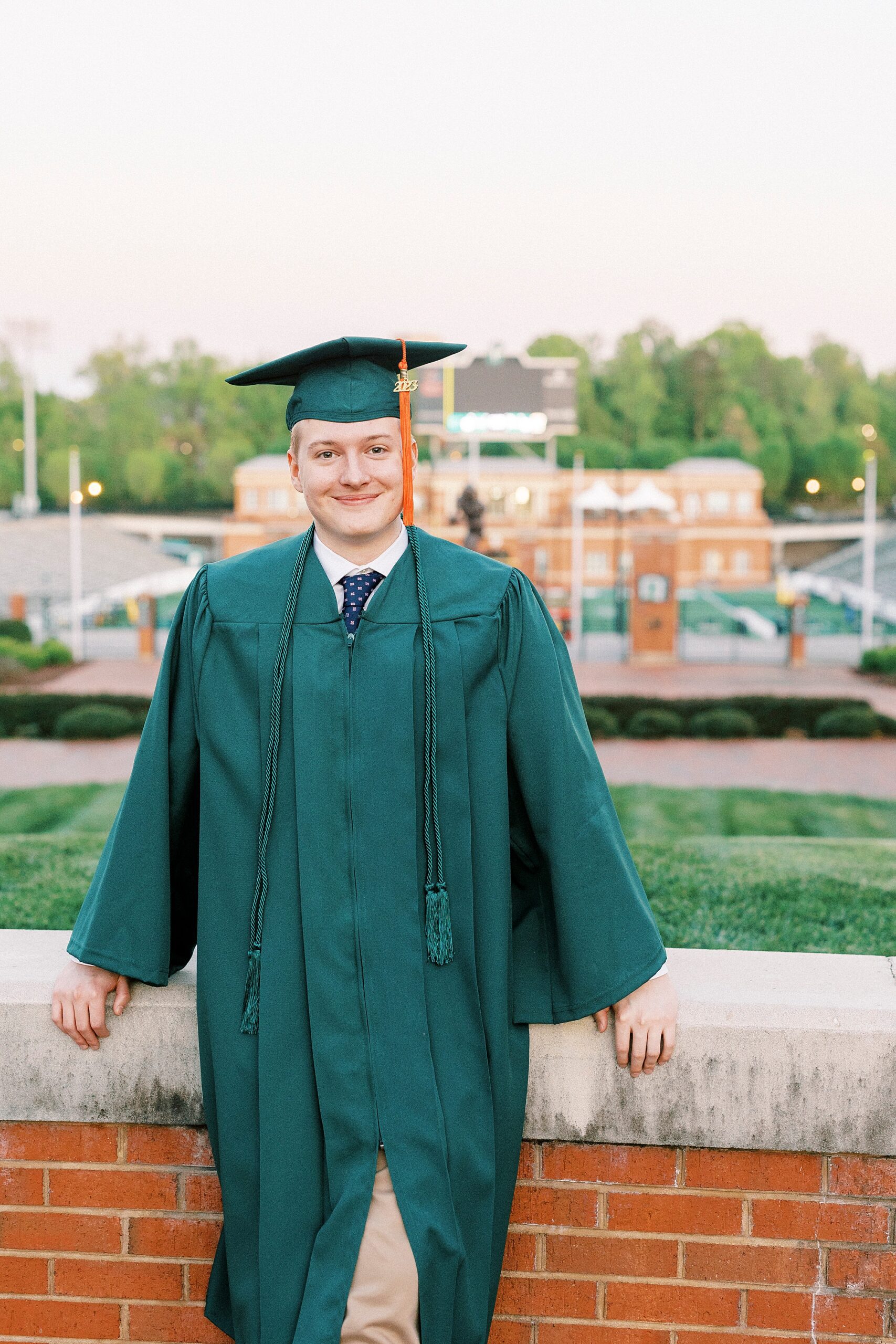 senior in green cap and gown leans against brick wall overlooking football field at UNC Charlotte