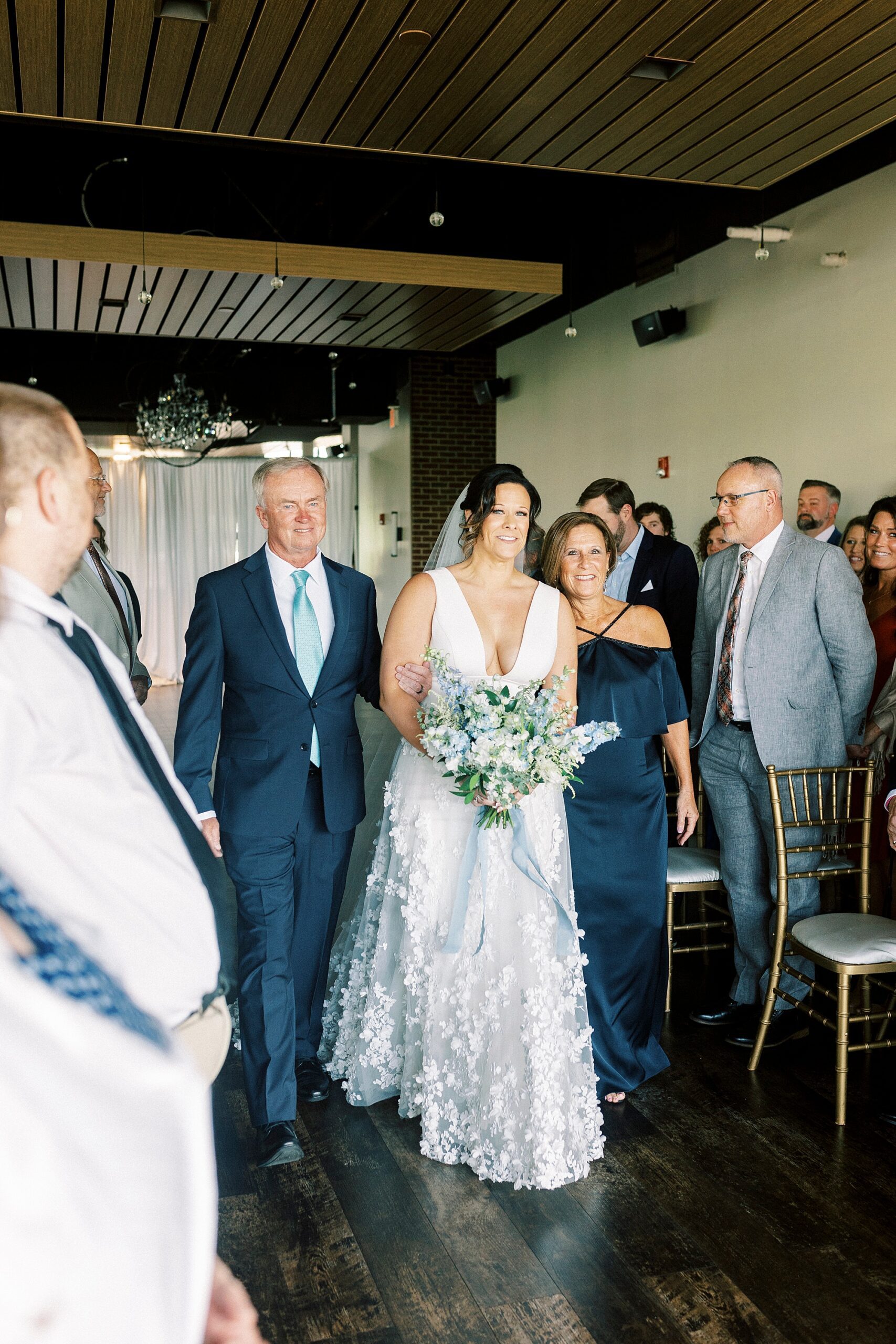 bride walks down aisle with father in blue suit carrying bouquet of blue and white flowers