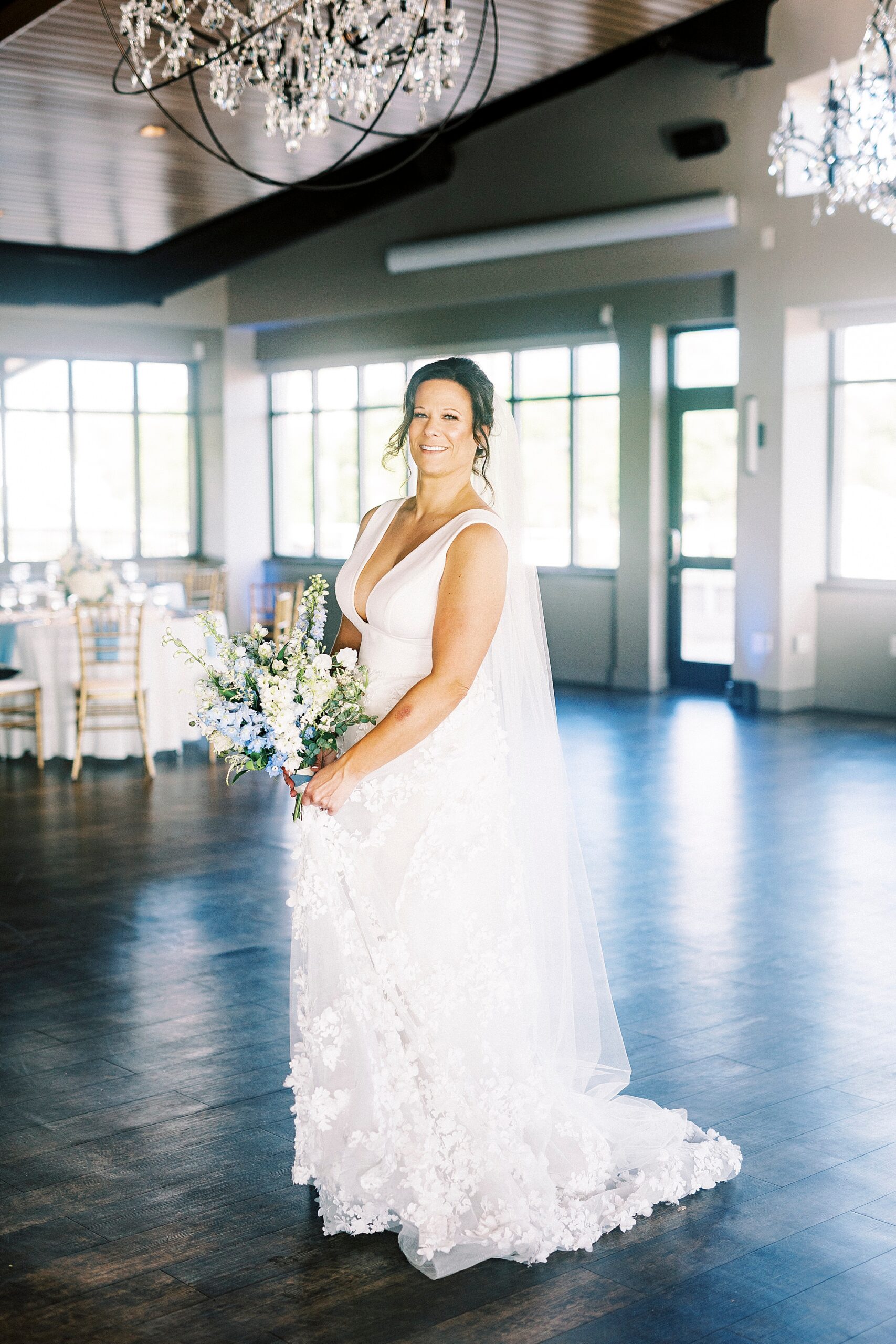 bride stands inside the Terrace at Cedar Hill near reception tables holding bouquet of blue and white flowers