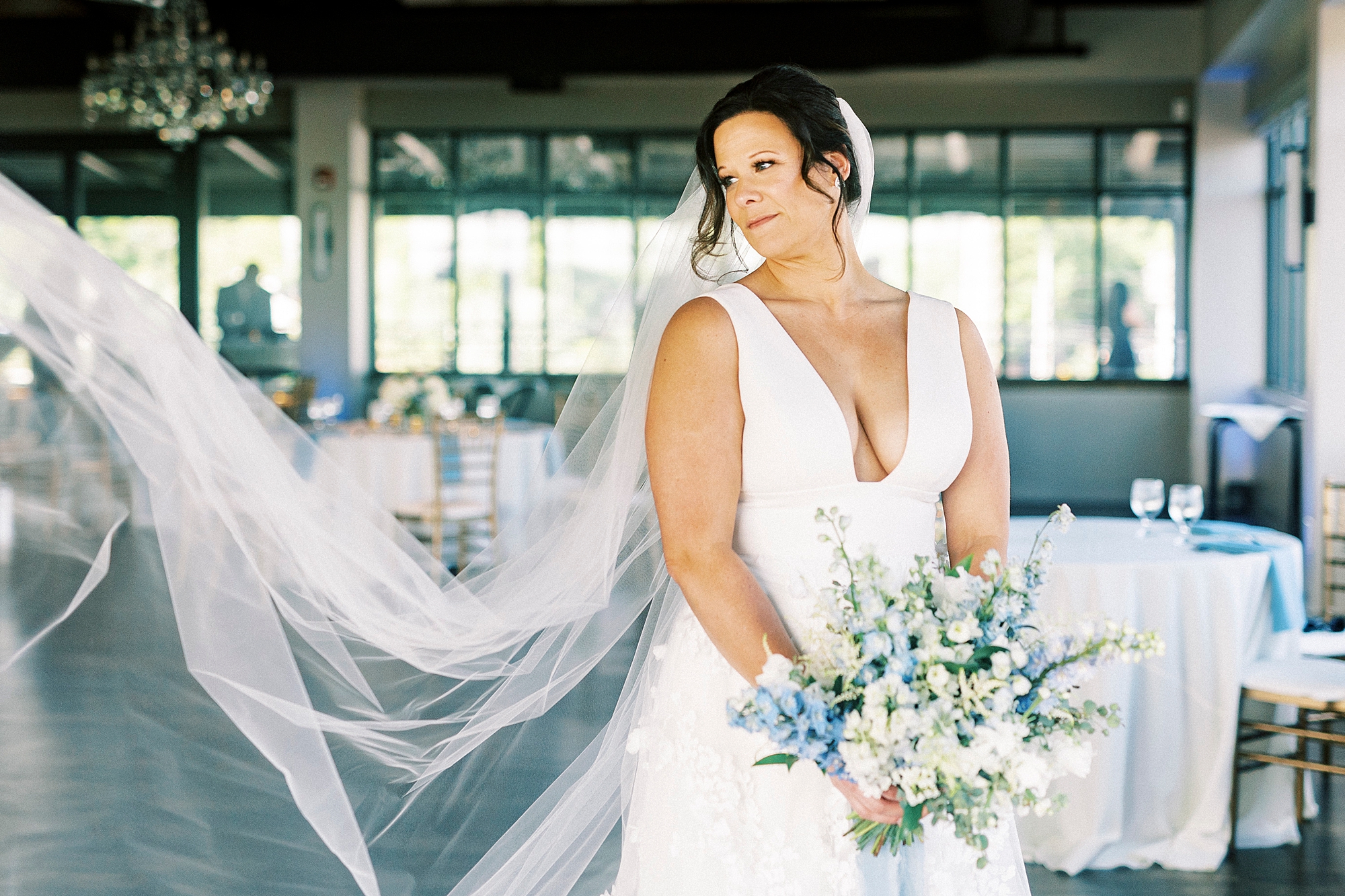 bride stands holding bouquet of blue and white flowers with veil floating