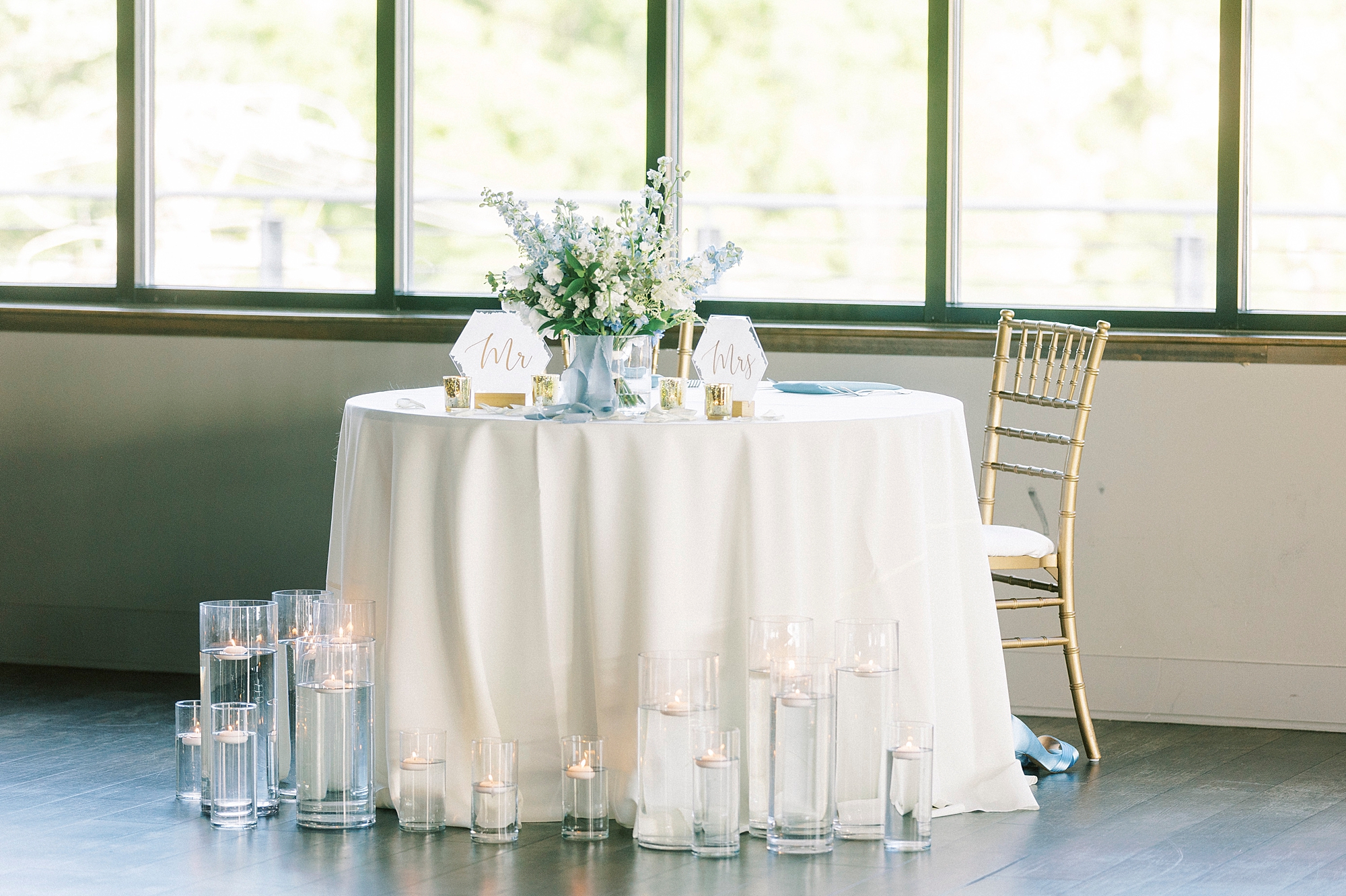 sweetheart table with white tablecloth, vases on floor with floating candles and blue and white bouquet on top