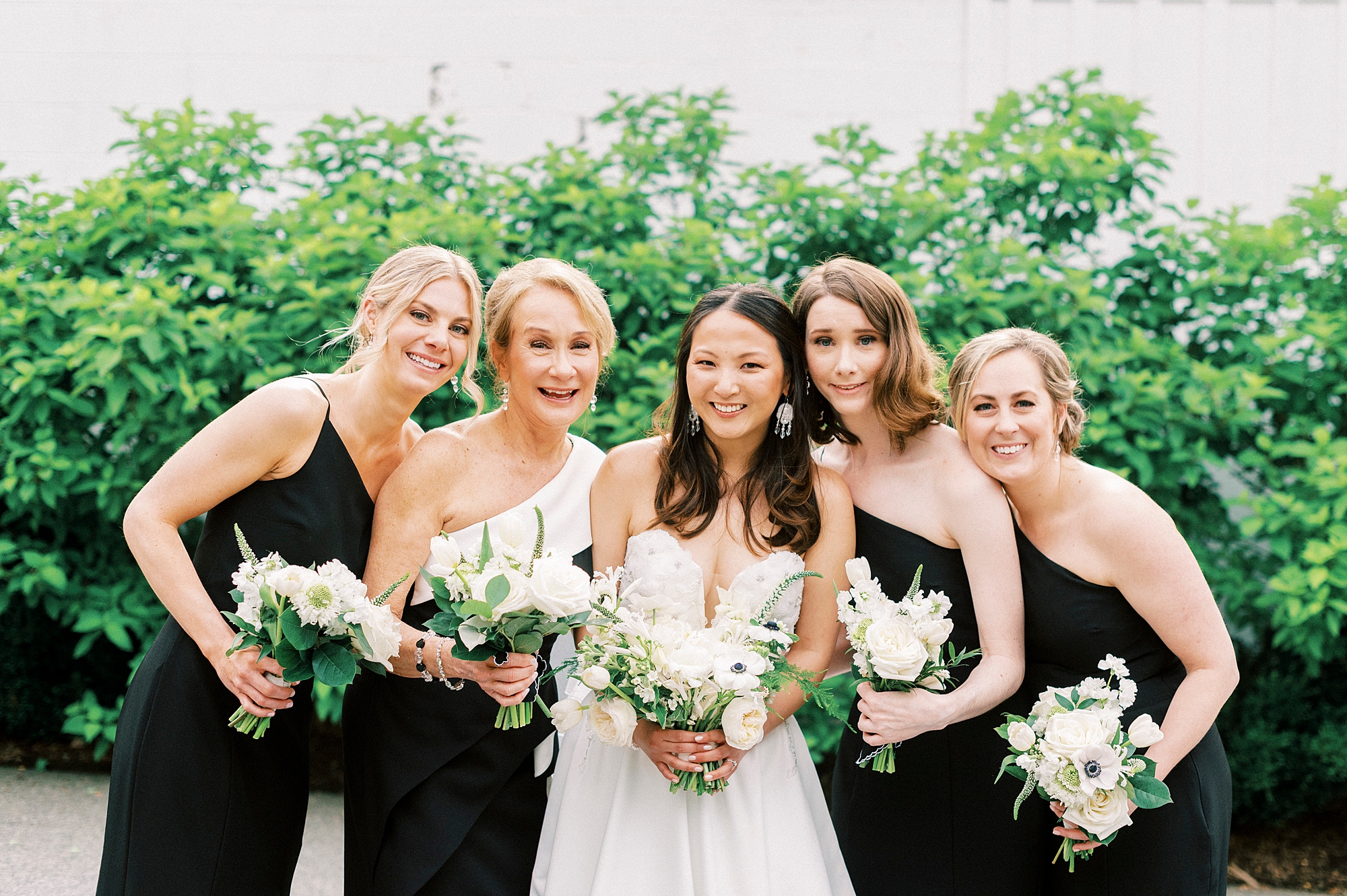 bride smiles with bridesmaids in one shoulder black gowns with ivory rose bouquets 