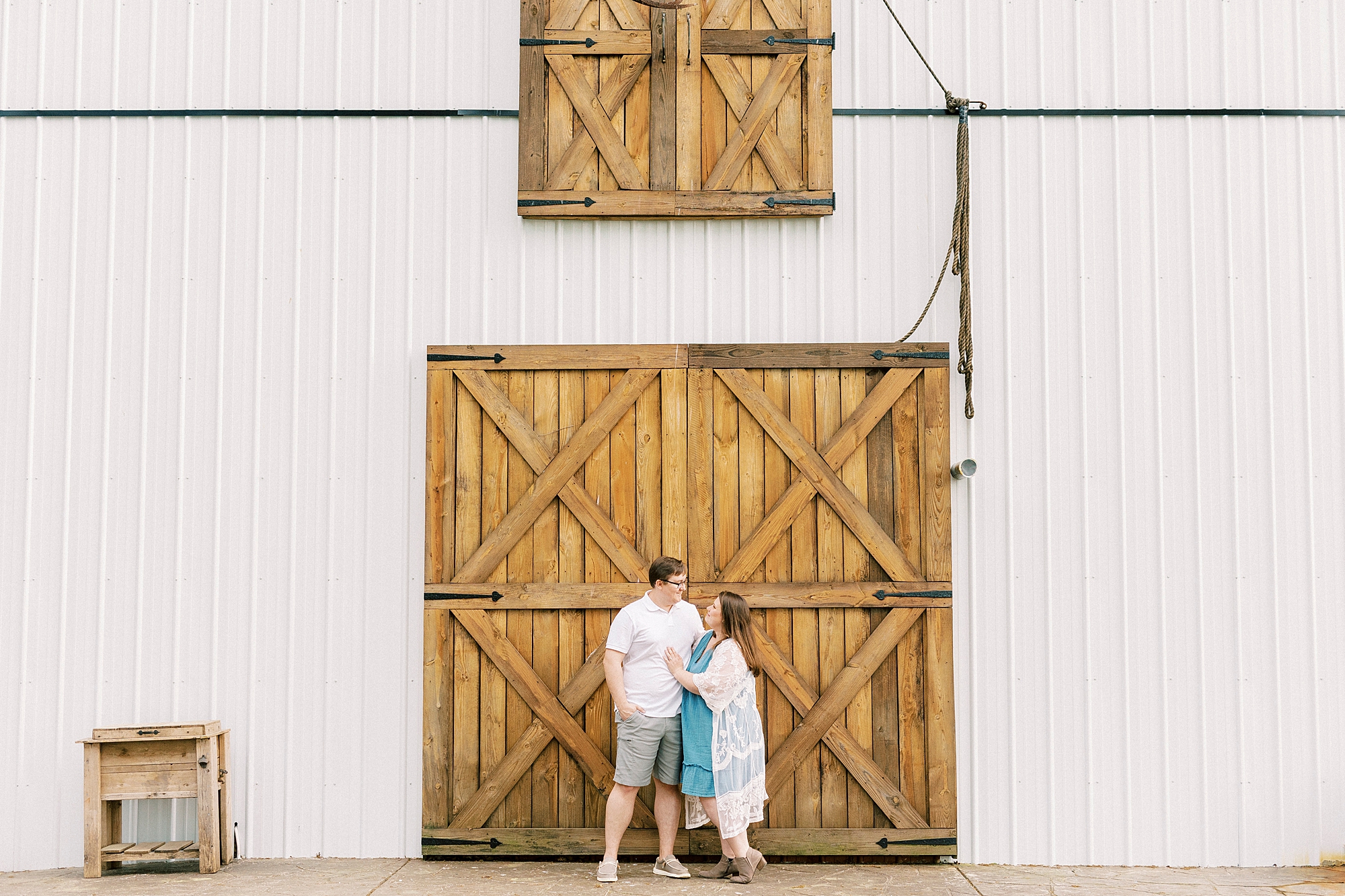 engaged couple hug in front of wooden doors on white barn at the Farmstead