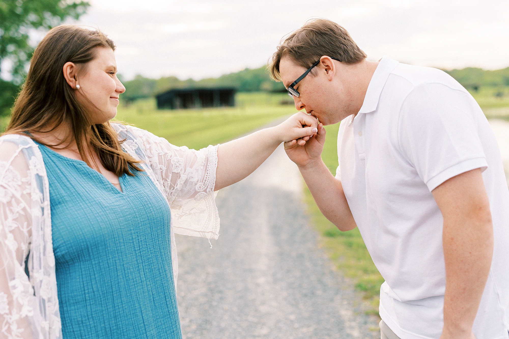 man in white polo kisses fiancee's hand on path at the Farmstead