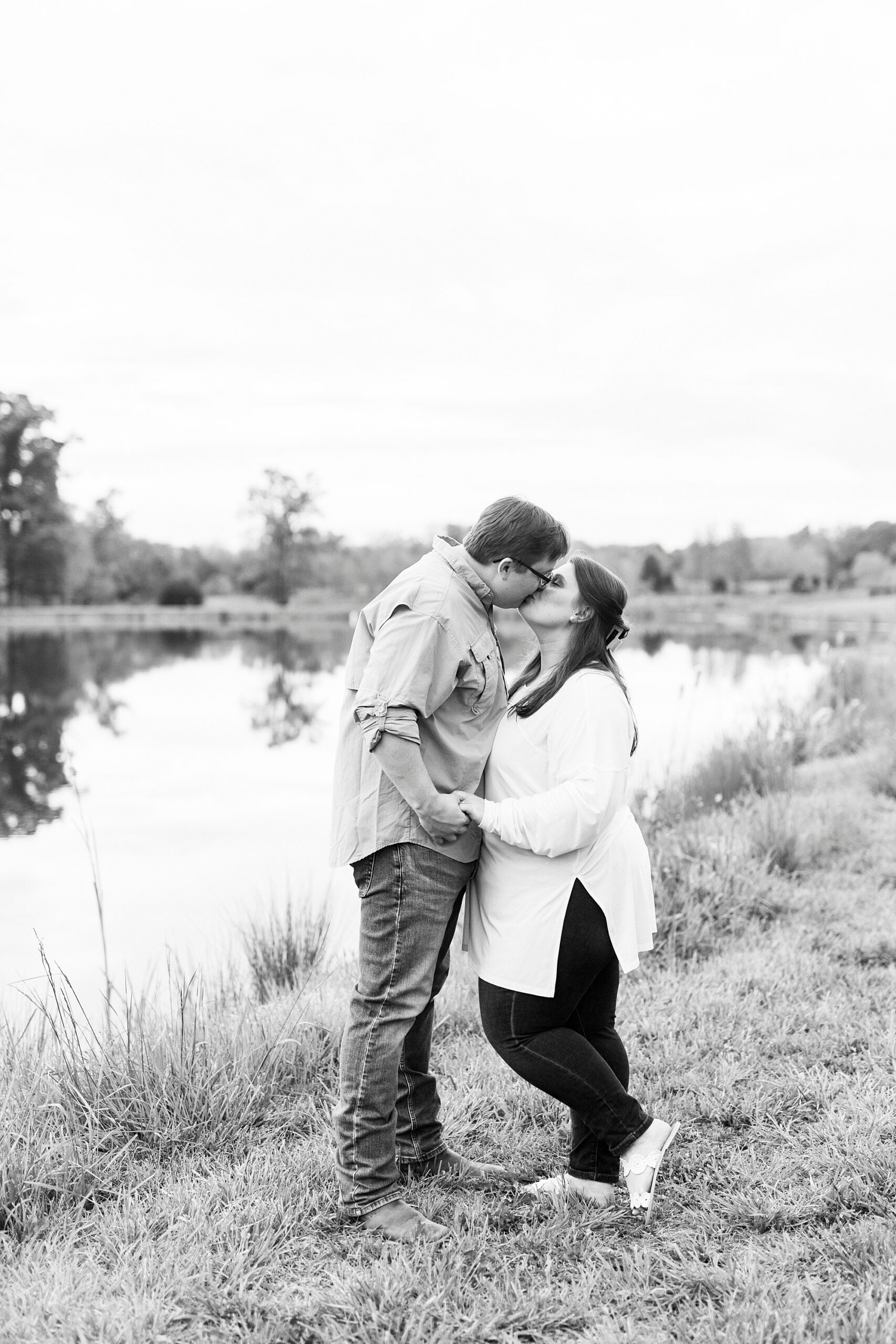 man leans to kiss fiancee in front of lake at the Farmstead
