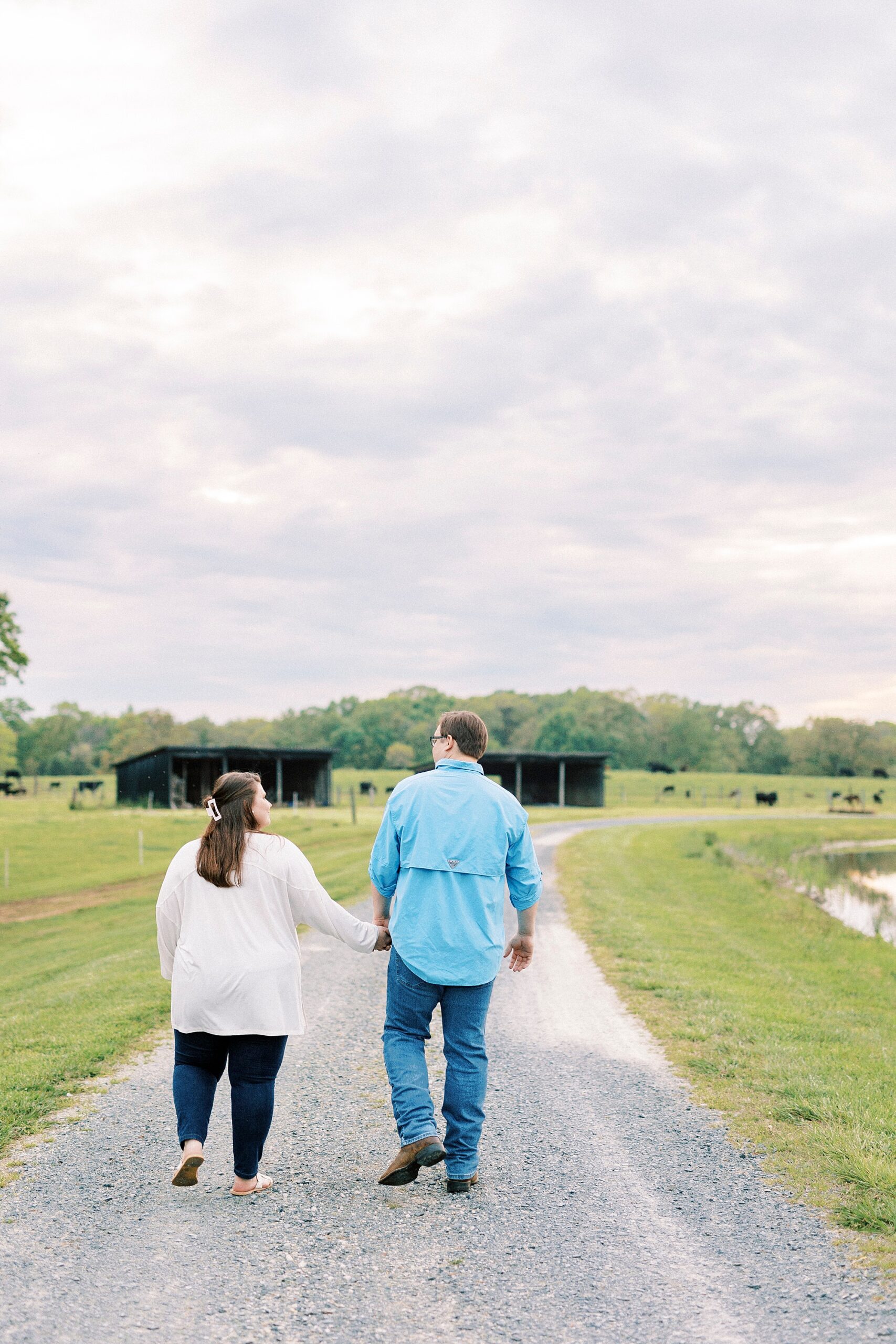 engaged couple holds hands walking on dirt path at the Farmstead