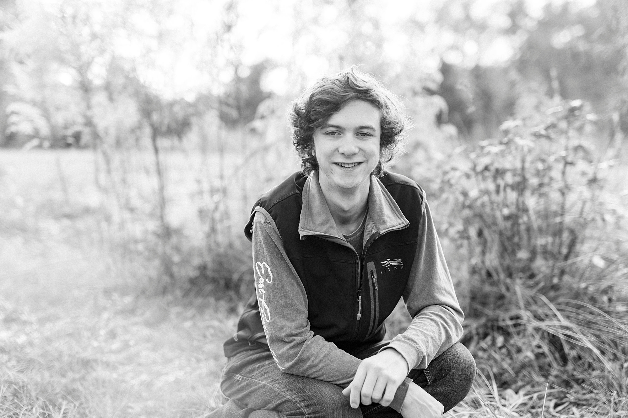 boy kneels down by tall grass during senior portraits in Midland NC