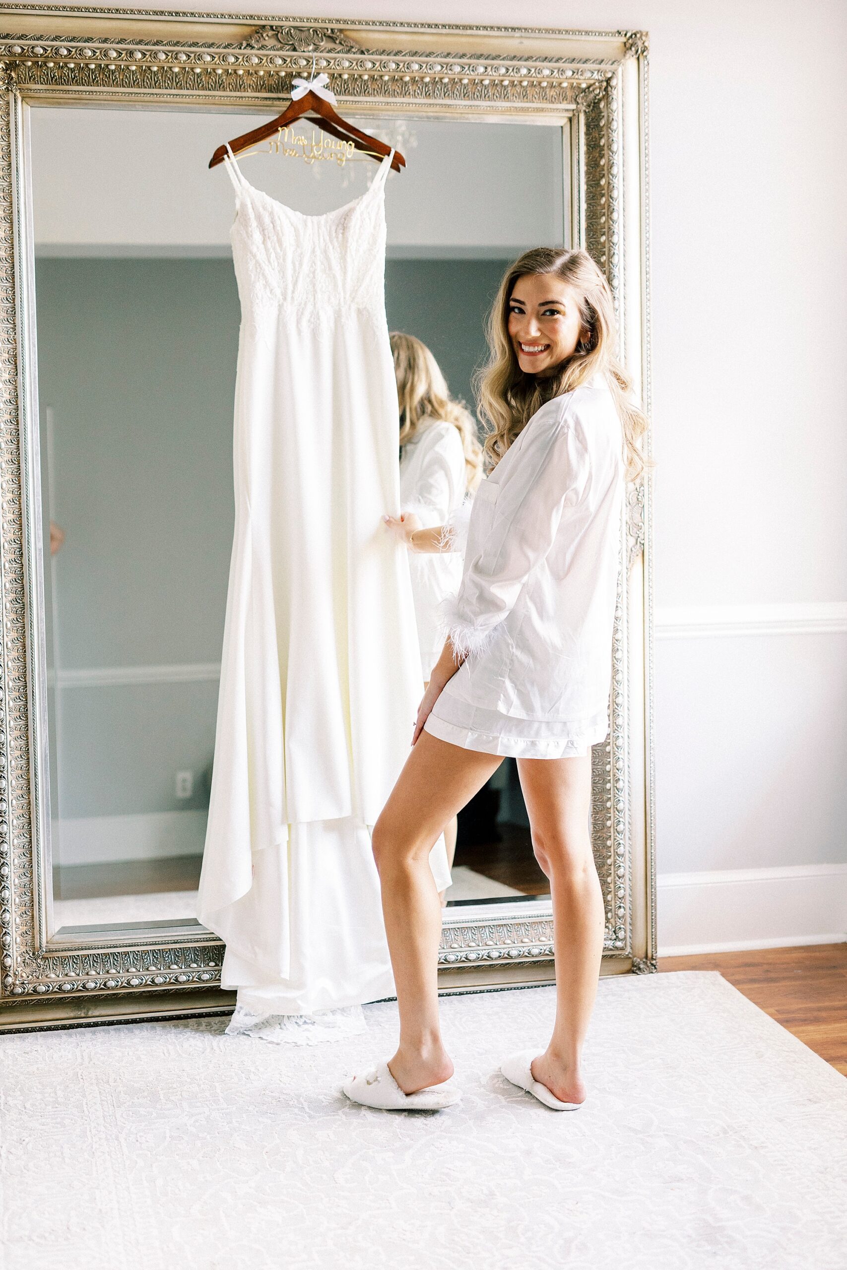 bride in white robe shows off wedding dress hanging on gold framed mirror 