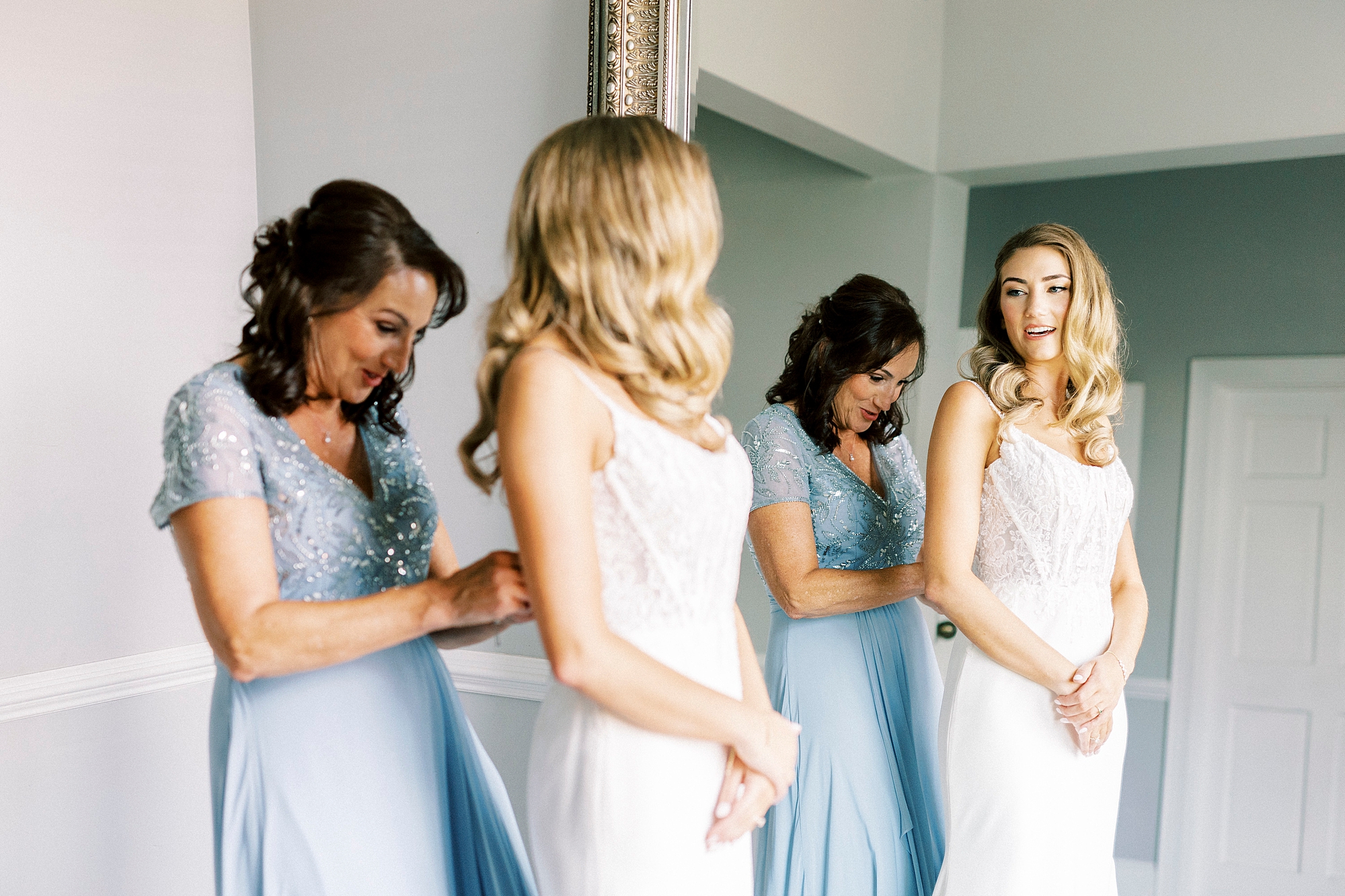 bridesmaid in light blue dress helps bride into wedding dress with reflection in mirror