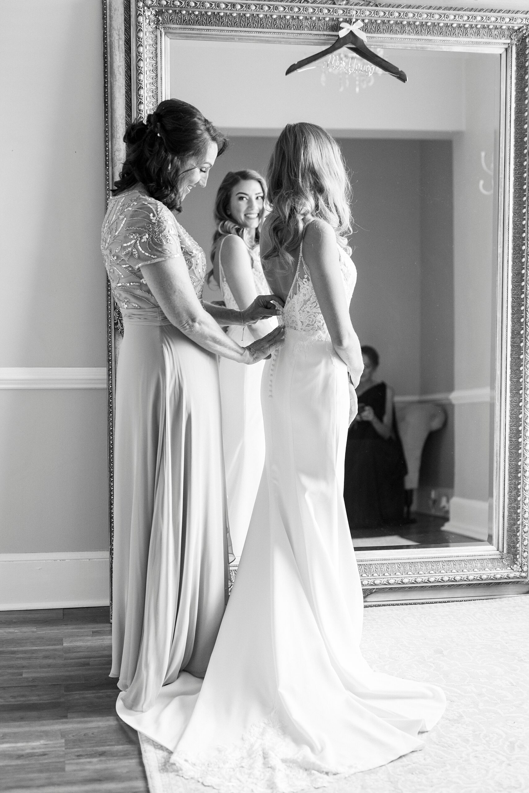 woman helps bride into wedding dress looking into mirror reflection 