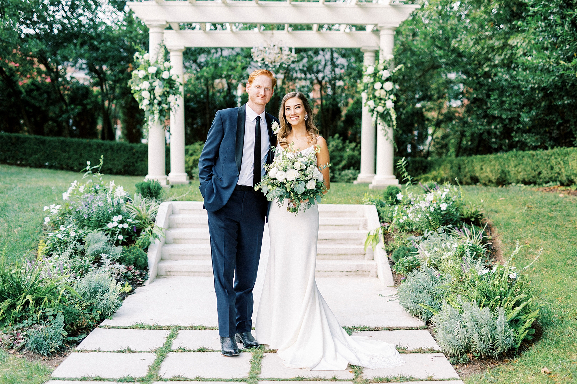 newlyweds hug in front of white arbor with greenery hanging behind them