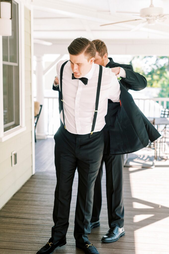 groomsman helps groom into black suit jacket on front porch at Whitehead Manor