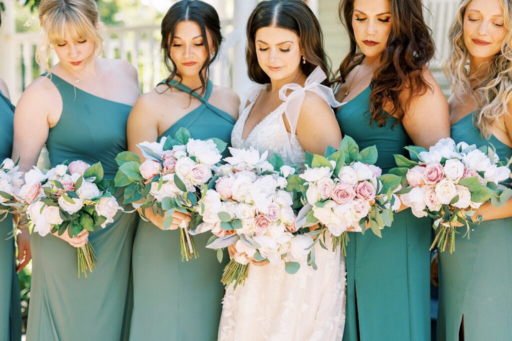 bride looks down at bouquets of pink and white flowers with bridesmaids in green dresses 
