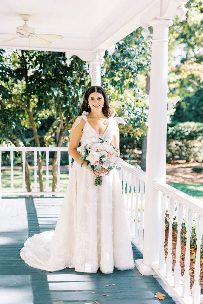 bride stands on front porch of Whitehead Manor with bouquet of white and pink flowers 