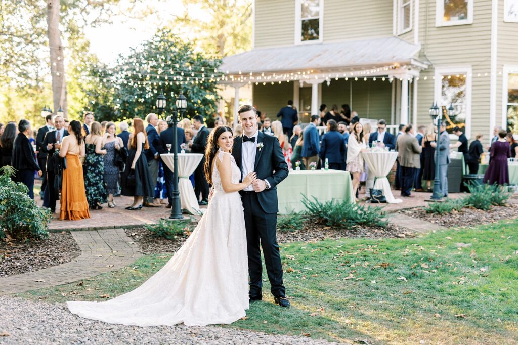 newlyweds stand on lawn with wedding guests behind them during cocktail hour 
