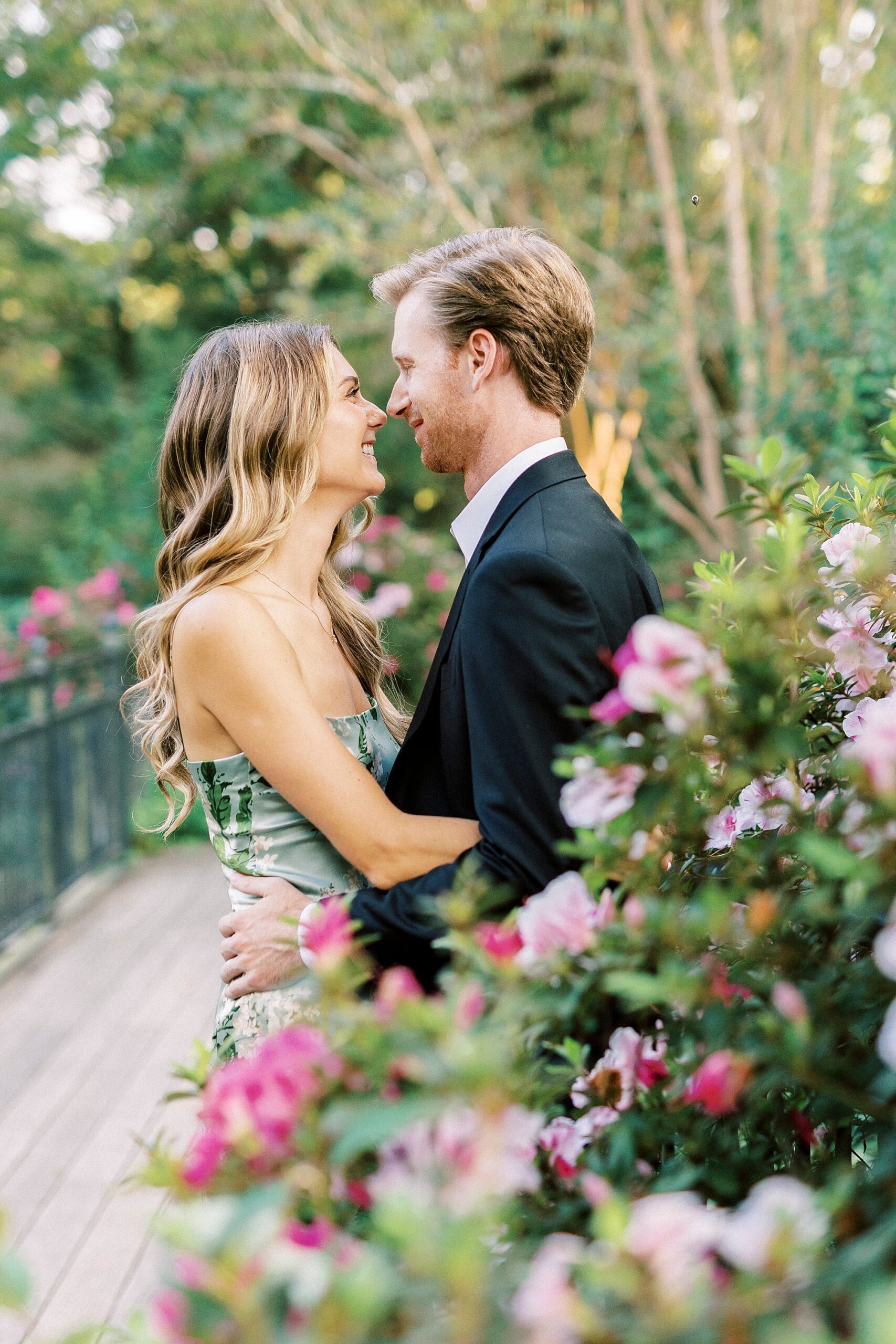 engaged couple hugs near pink flowers on bridge at Glencairn Garden