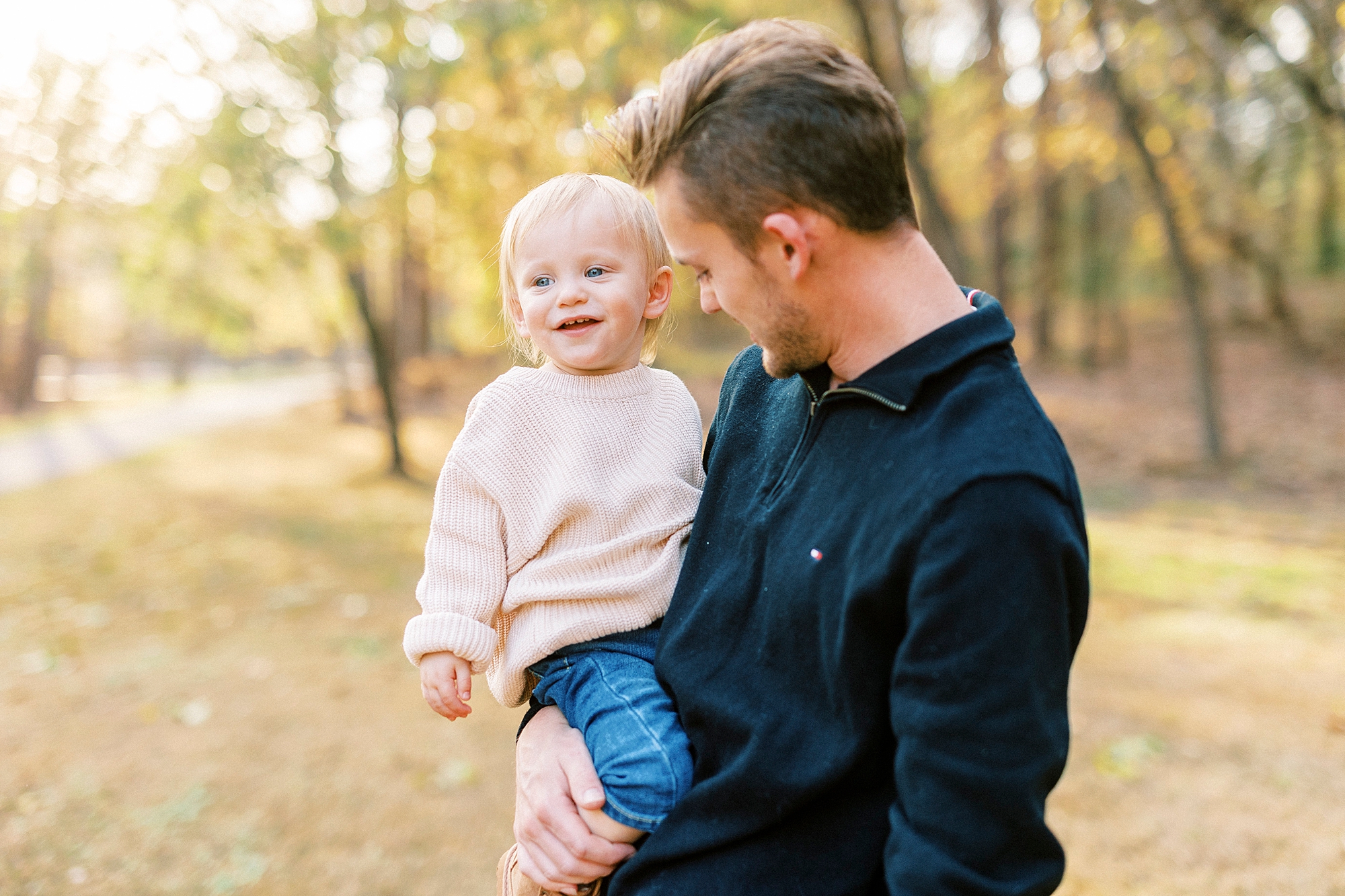 dad in black shirt looks at toddler in cream sweater during fall family photos 