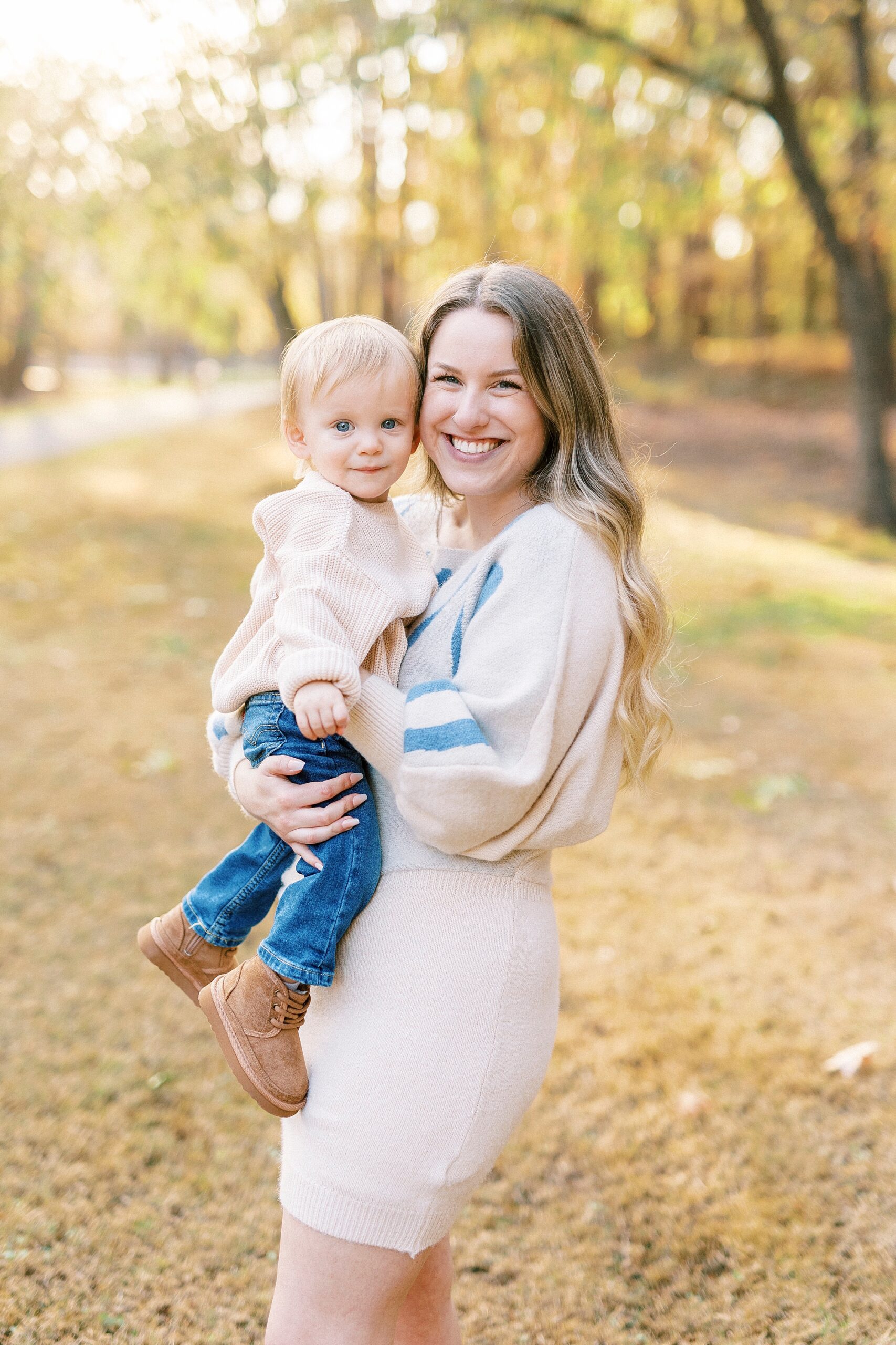 blonde woman in cream sweater dress smiles holding son to her