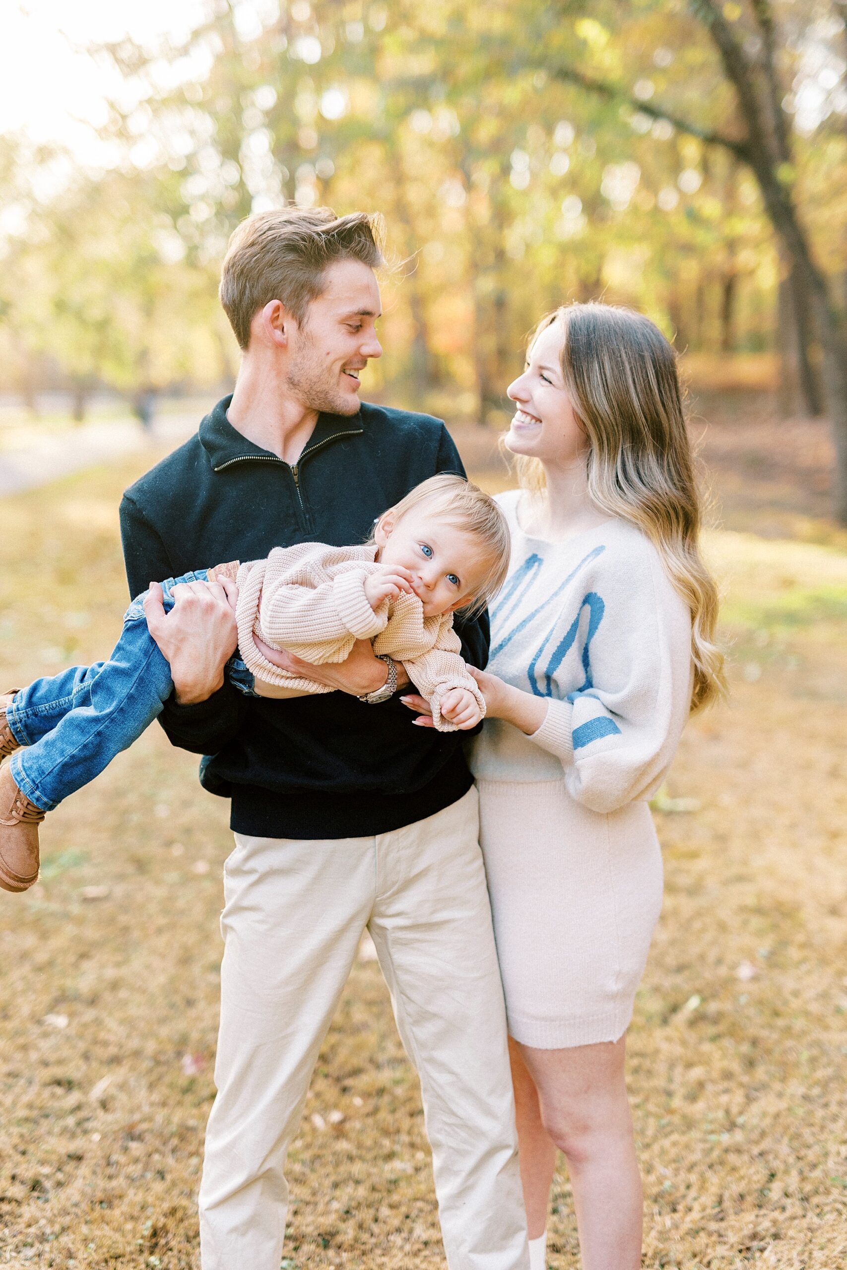 couple smiles while son lays in their arms during fall family portraits 