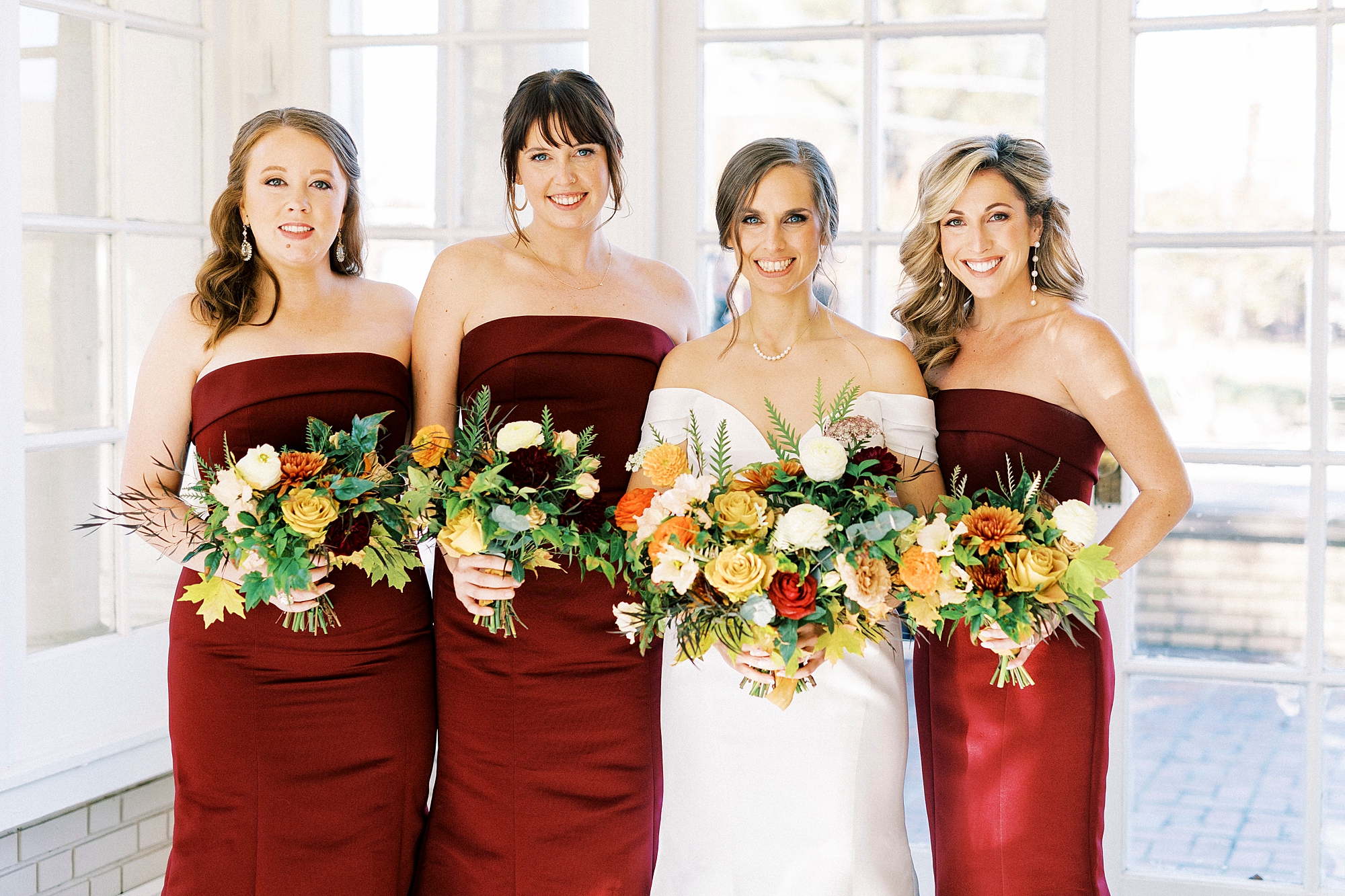 bride stands with bridesmaids in burgundy gowns with orange and yellow flowers 