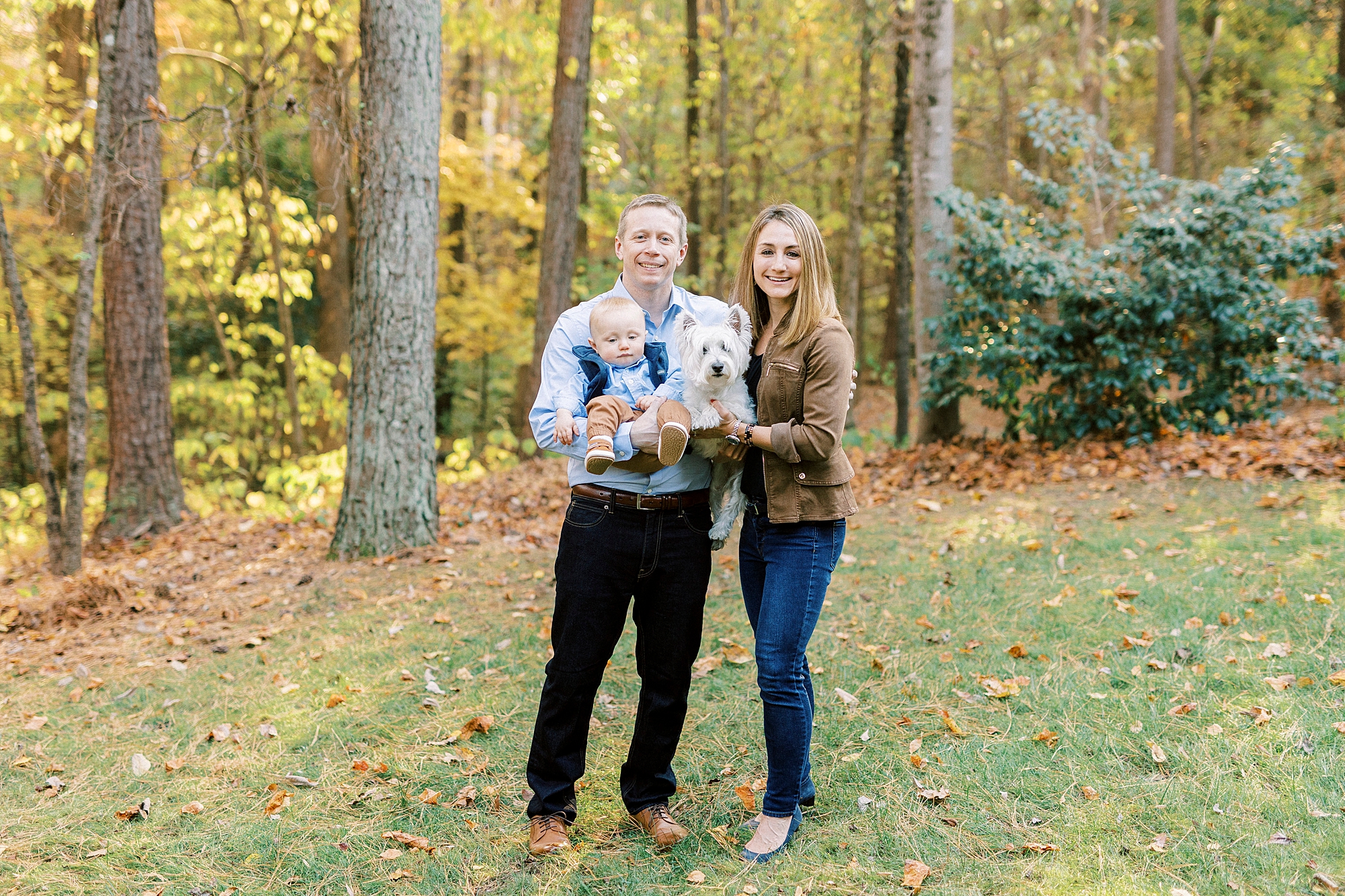 parents pose with son and dog in their arms in front of woods in backyard 