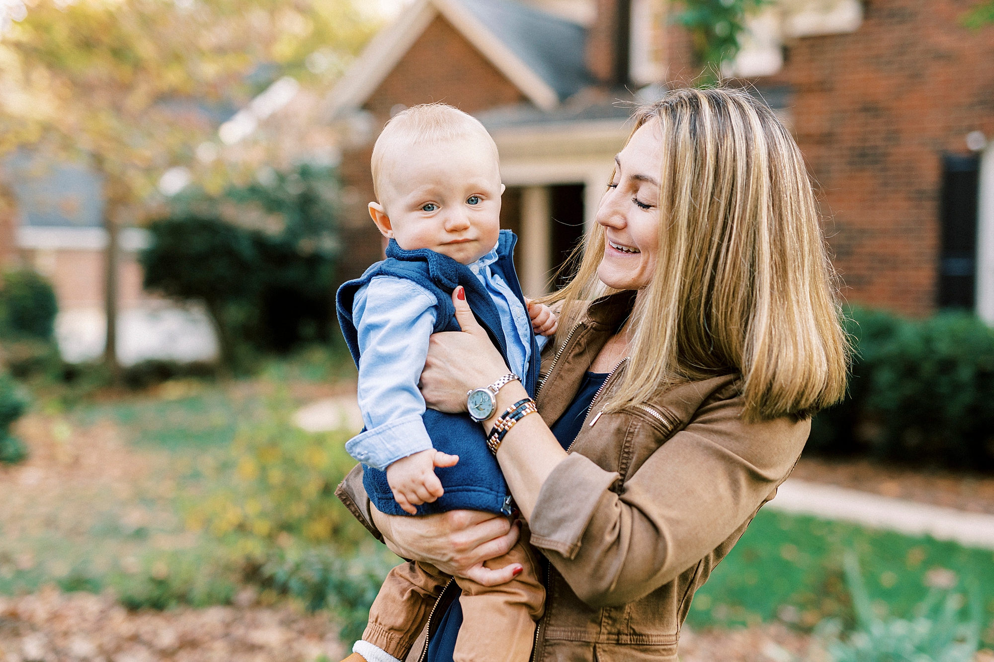 mom in brown jacket holds son in blue vest during fall family portraits at Charlotte home