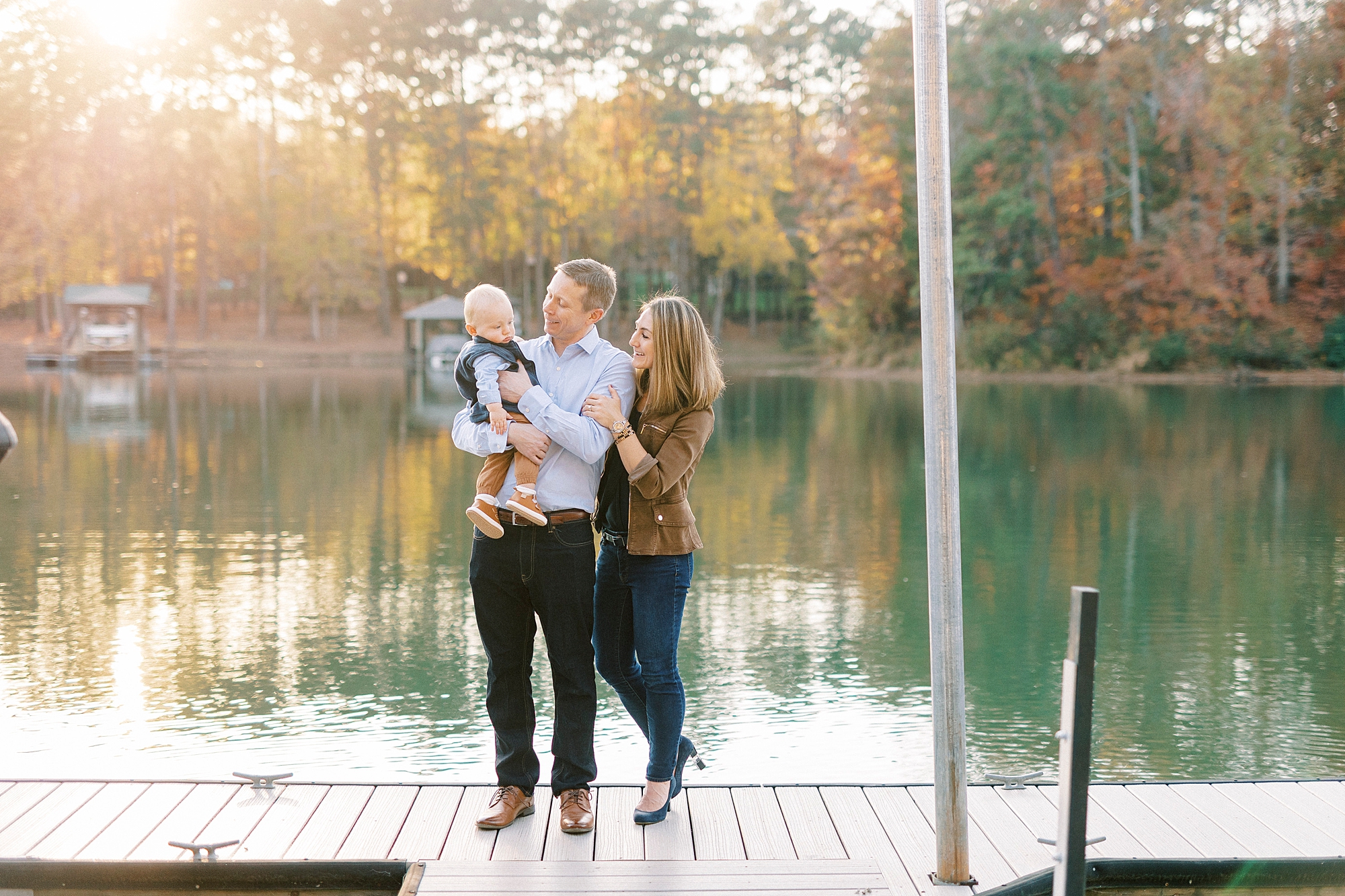 parents smile at son on dad's hip during fall family portraits at Charlotte home on lake