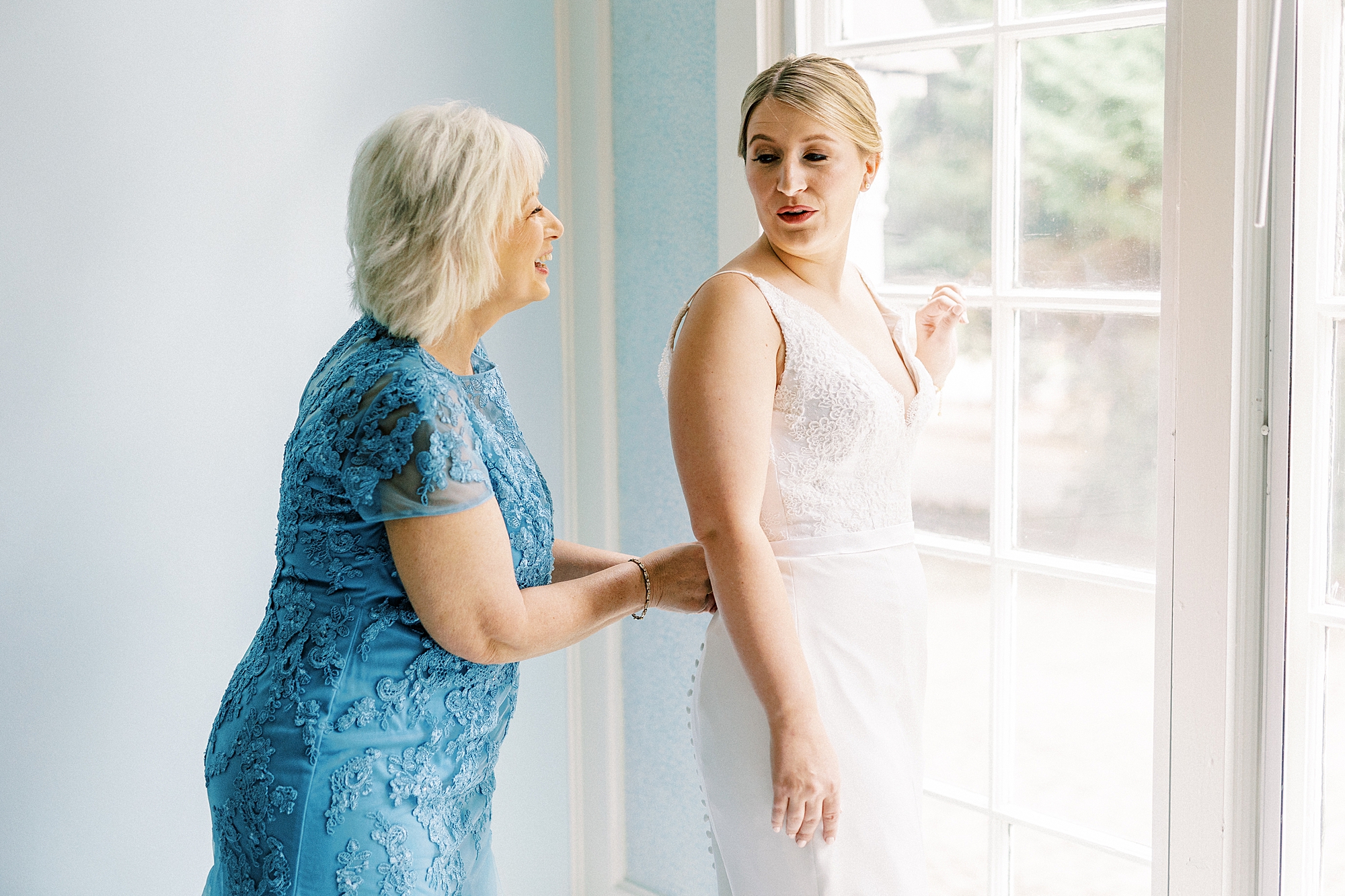 mother in blue dress buttons up wedding gown for bride looking out window 