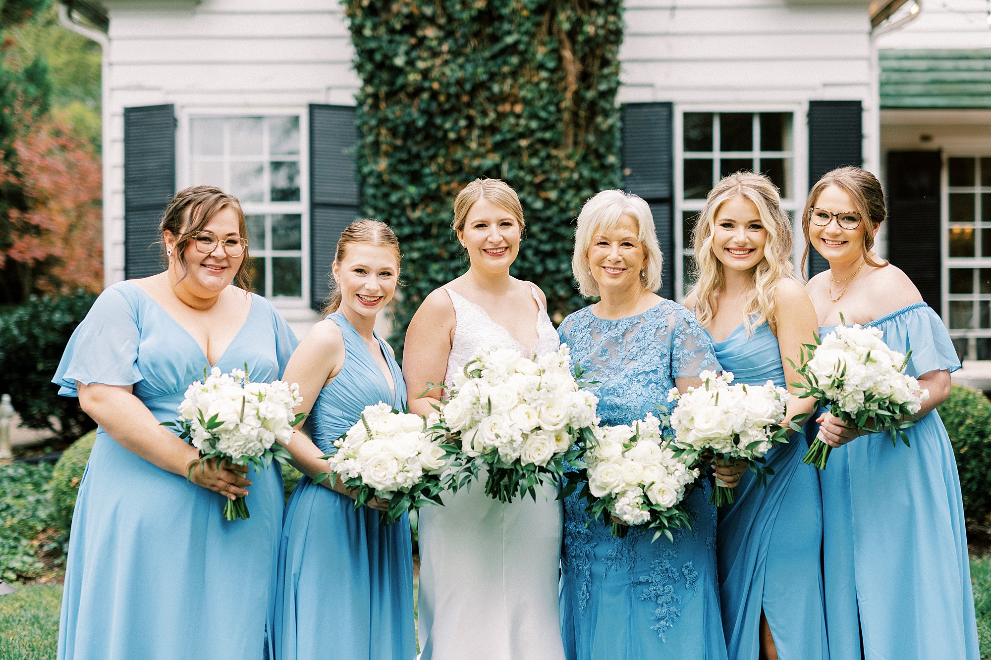 bride poses with bridesmaids in light blue gowns with white bouquets outside the Morehead Inn