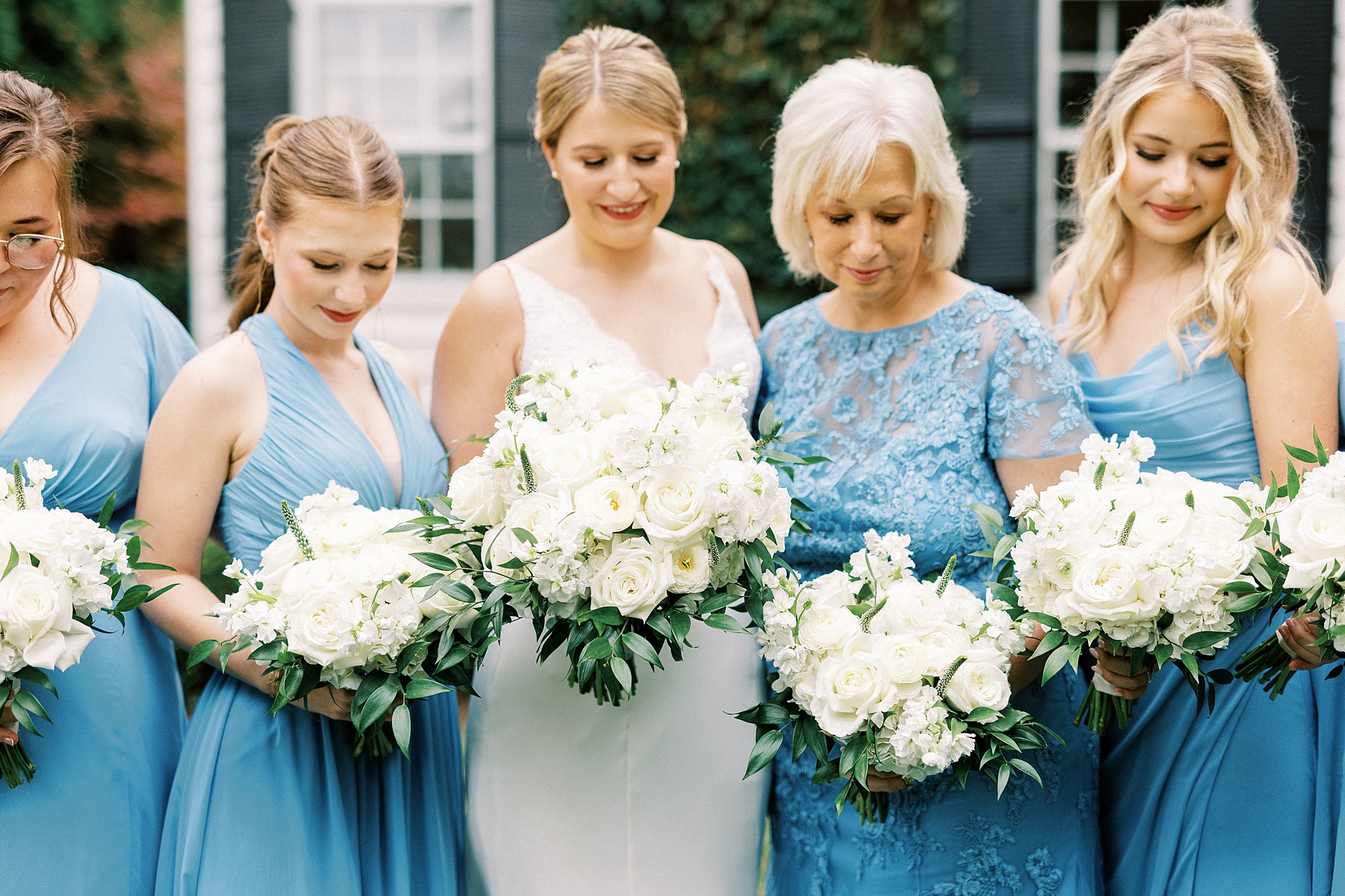 bride looks down at bouquet of ivory flowers surrounded by bridesmaids in light blue gowns 