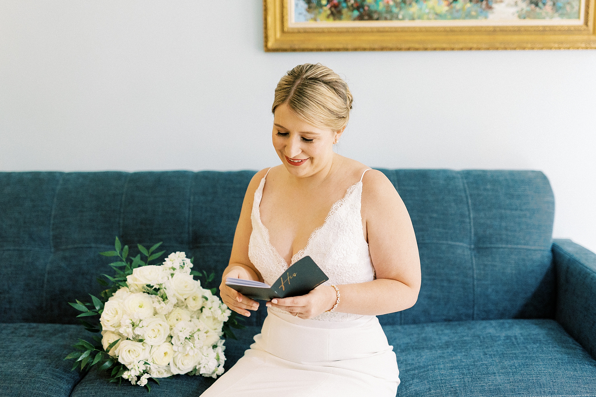bride sits on blue couch reading vow booklet on morning of wedding