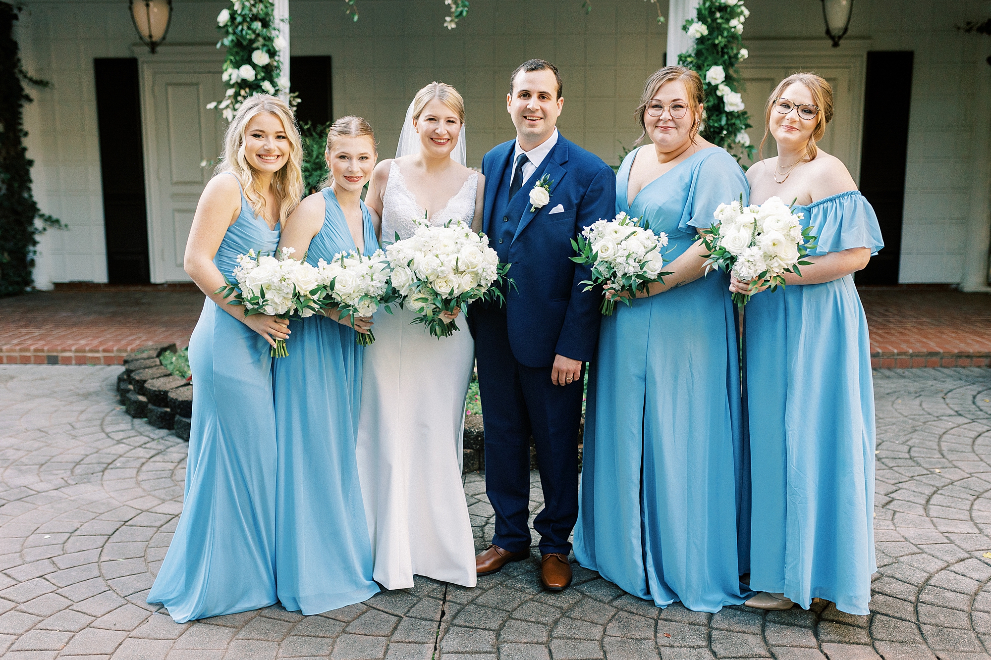 bride and groom stands with bridesmaids in blue gowns in front of arbor at the Morehead Inn