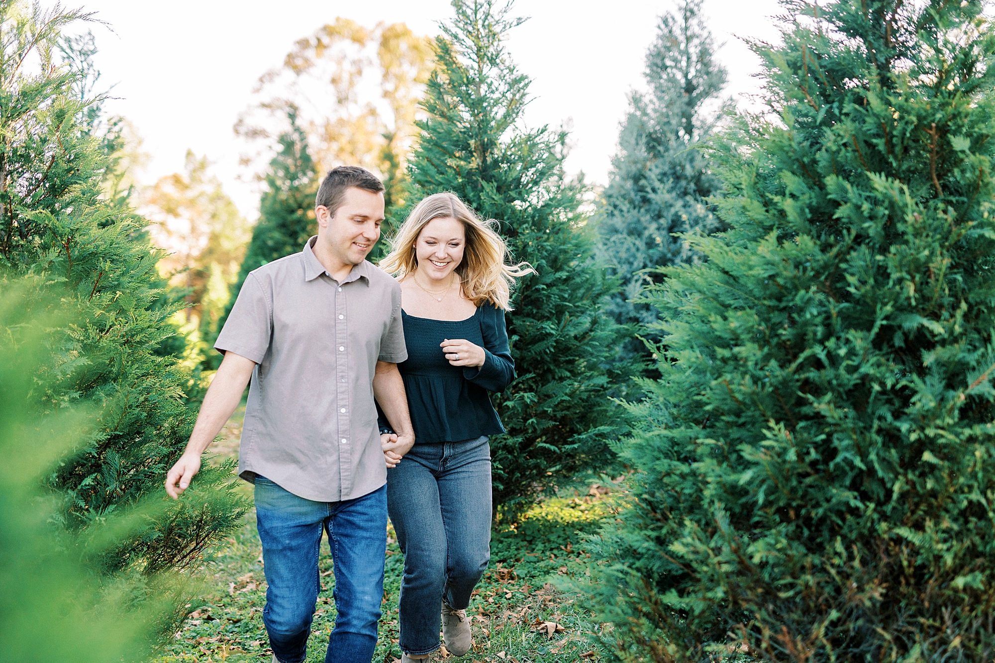 couple holds hands running between trees at Almond Christmas Tree Farm