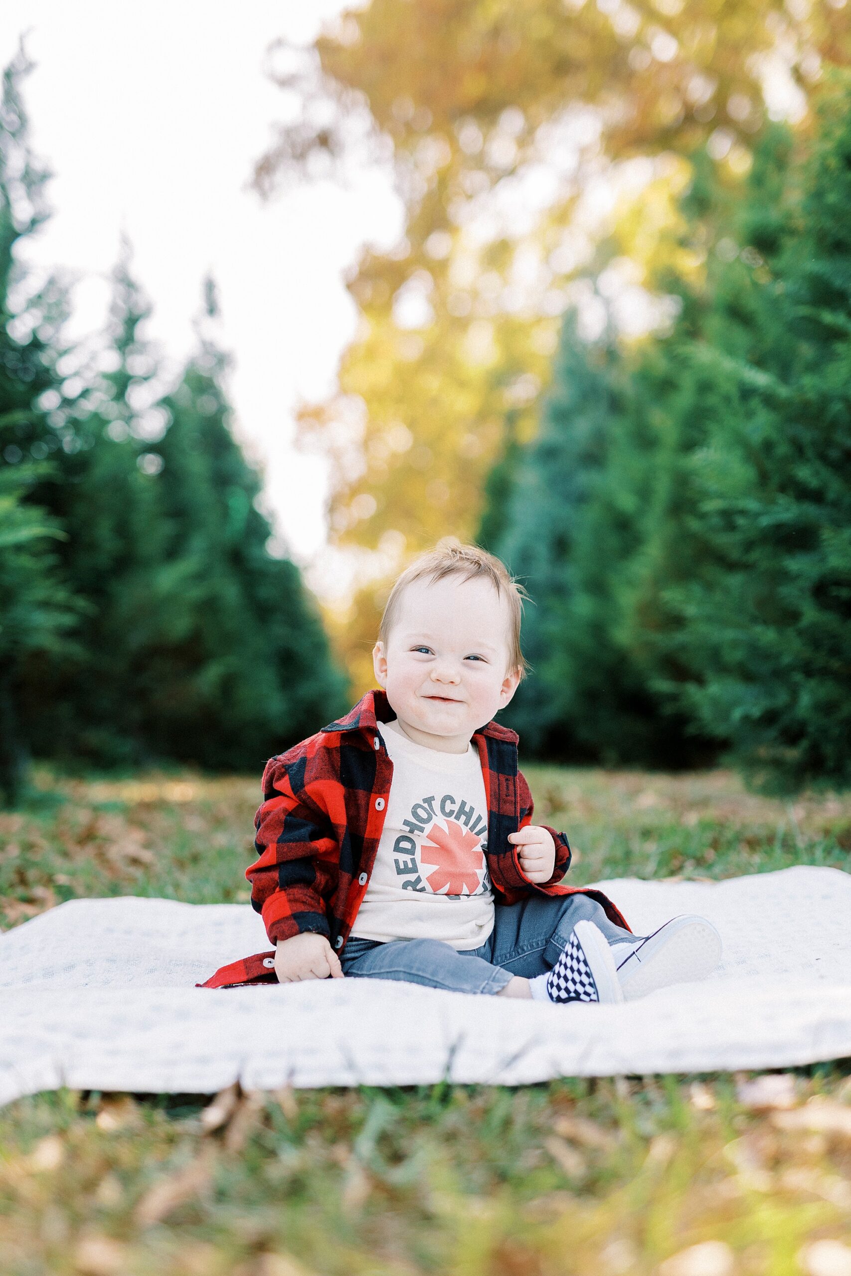 baby in buffalo plaid shirt sits on blanket in front of Christmas trees