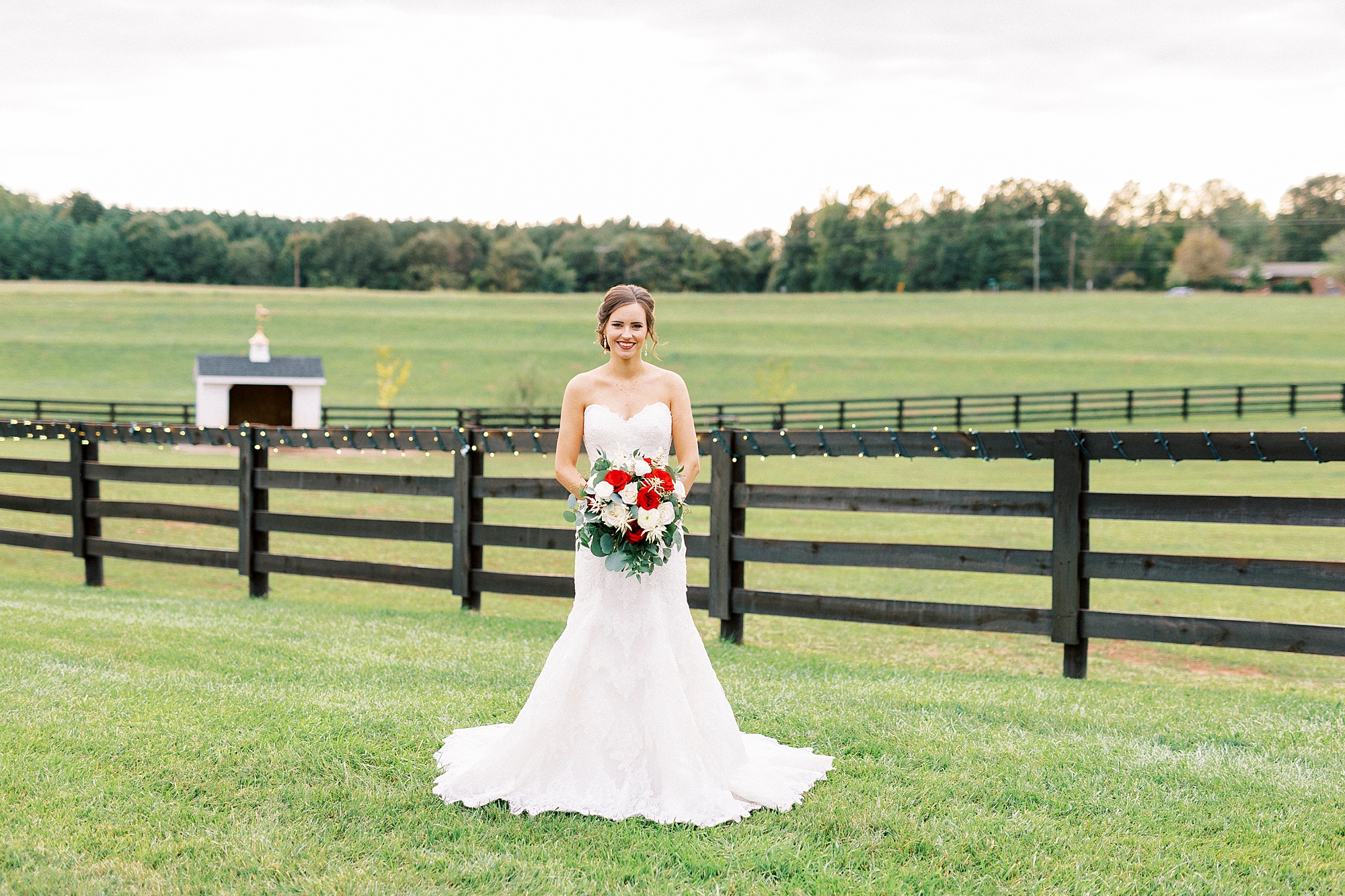 bride stands in front of wooden fence at Chickadee Hill Farms holding bouquet of red and white flowers 