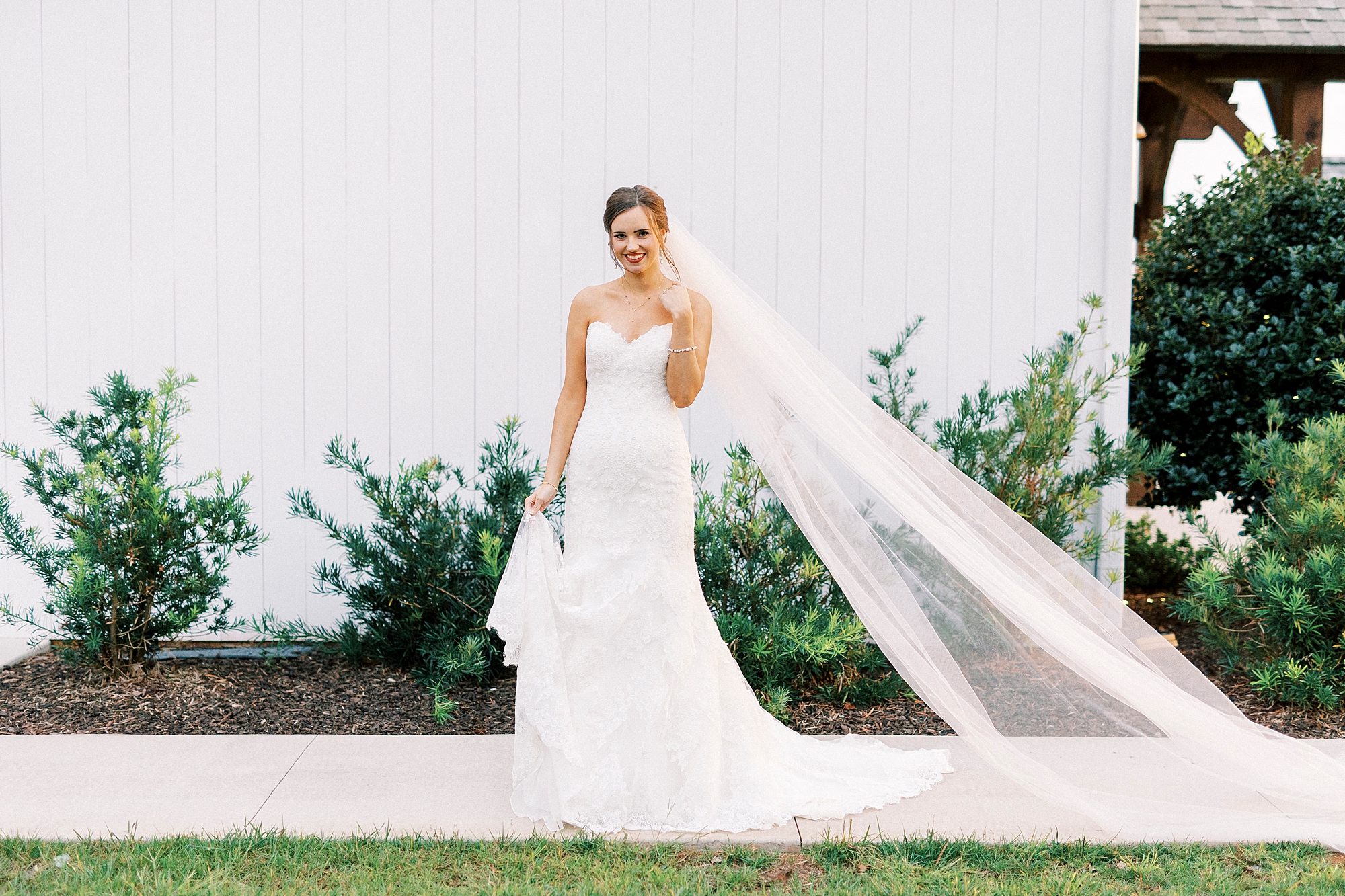 bride stands with veil dragging behind her outside white wall of Chickadee Hill Farms chapel 