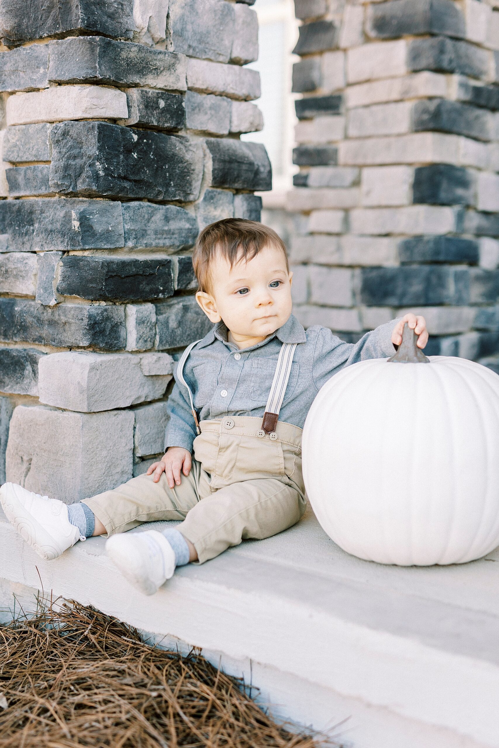boy in blue shirt in suspenders sits next to white pumpkin on front porch 