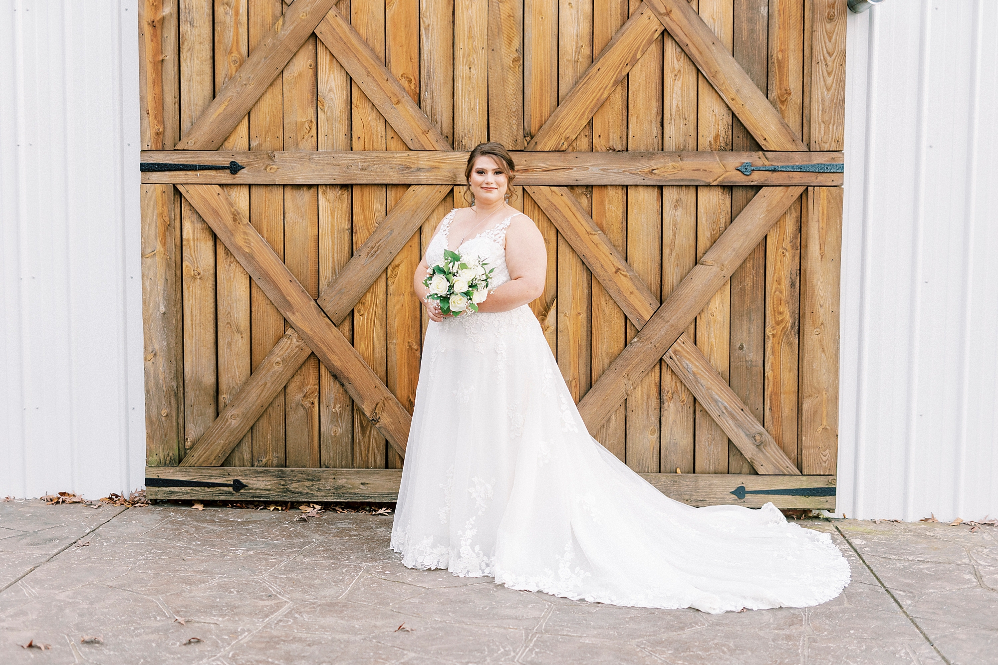 bride stands holding bouquet of white roses and baby's breath in front of wooden doors 