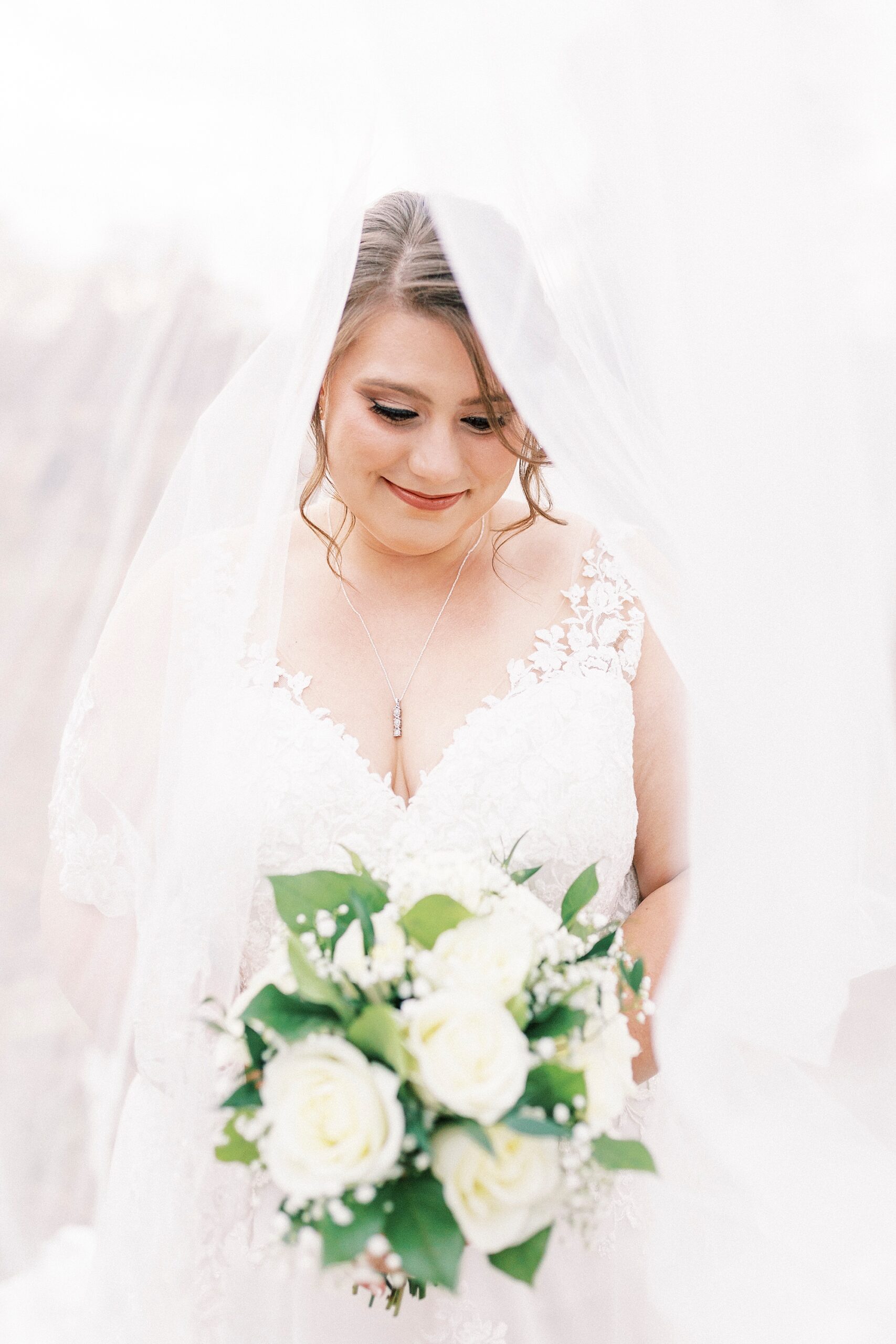 bride looks down at bouquet of white roses and baby's breath with veil pulled over her head